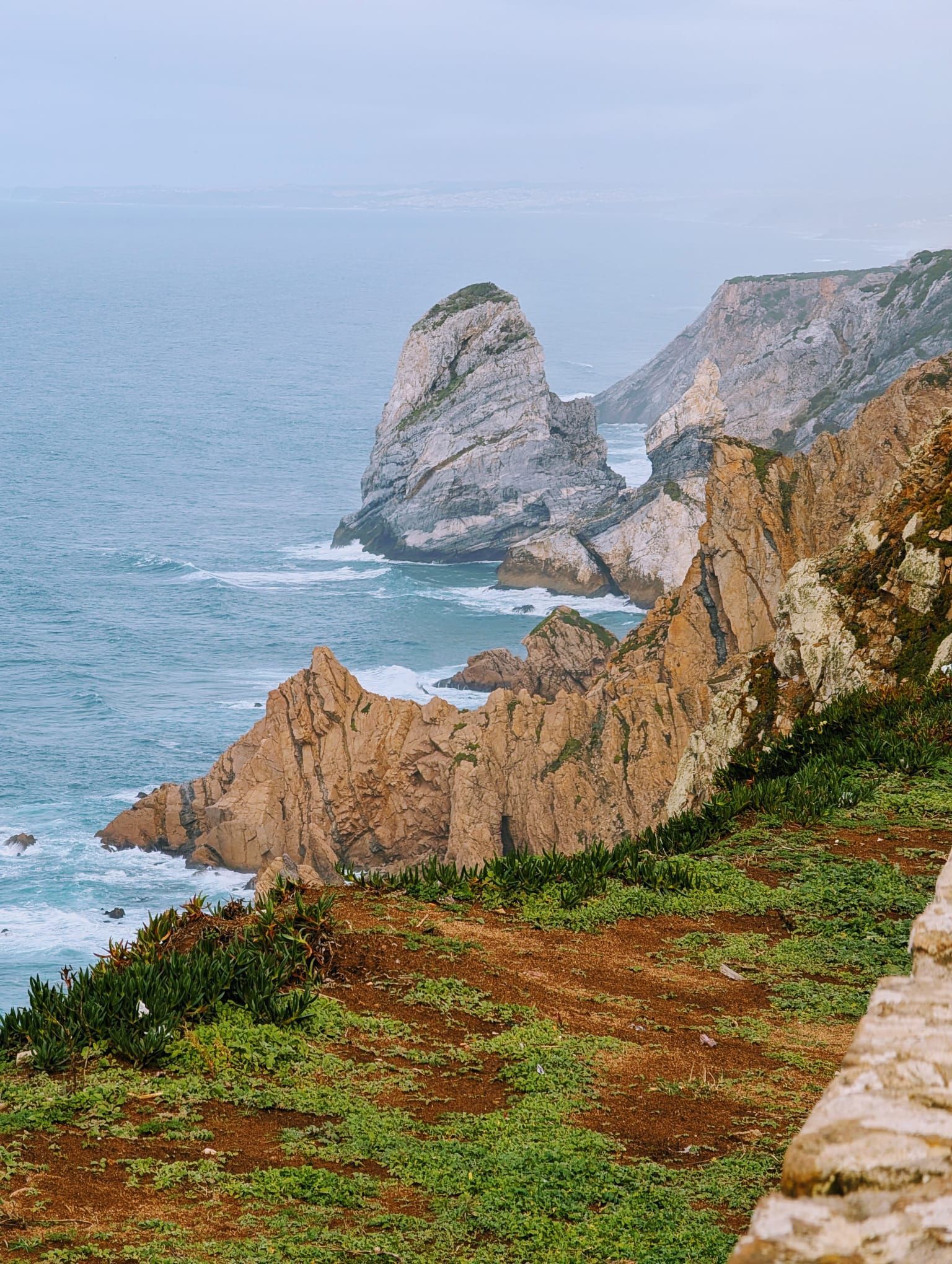 Wooden walkway over the sand at Costa Nova Beach, with the calm sea to the side and, in the distance, the Barra Lighthouse — one of the tallest in Europe — under a partly cloudy evening sky.