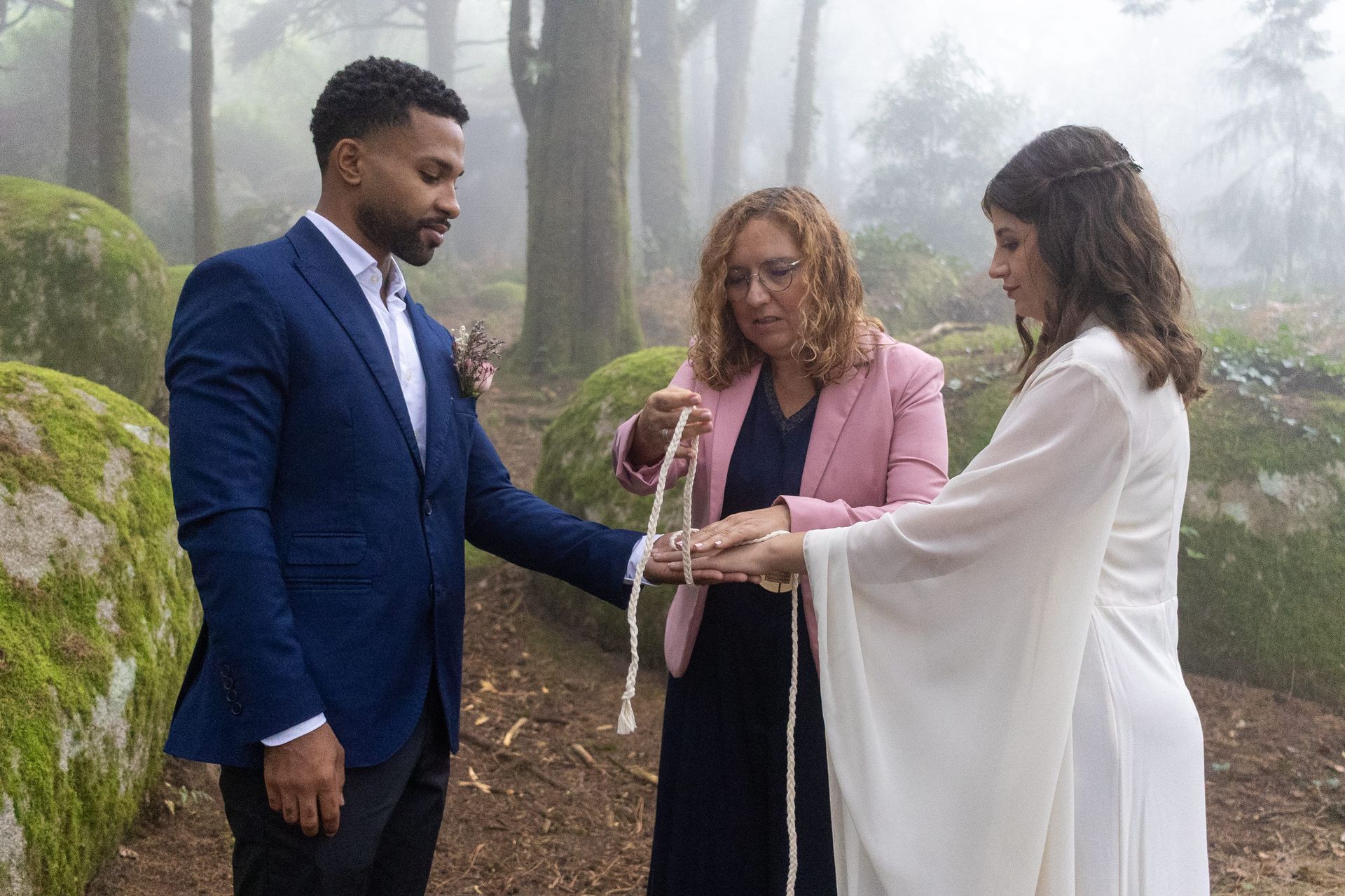 A handfasting ritual in the foggy forest of Sintra. A couple and an officiant are present, with a white cord being tied around their hands.