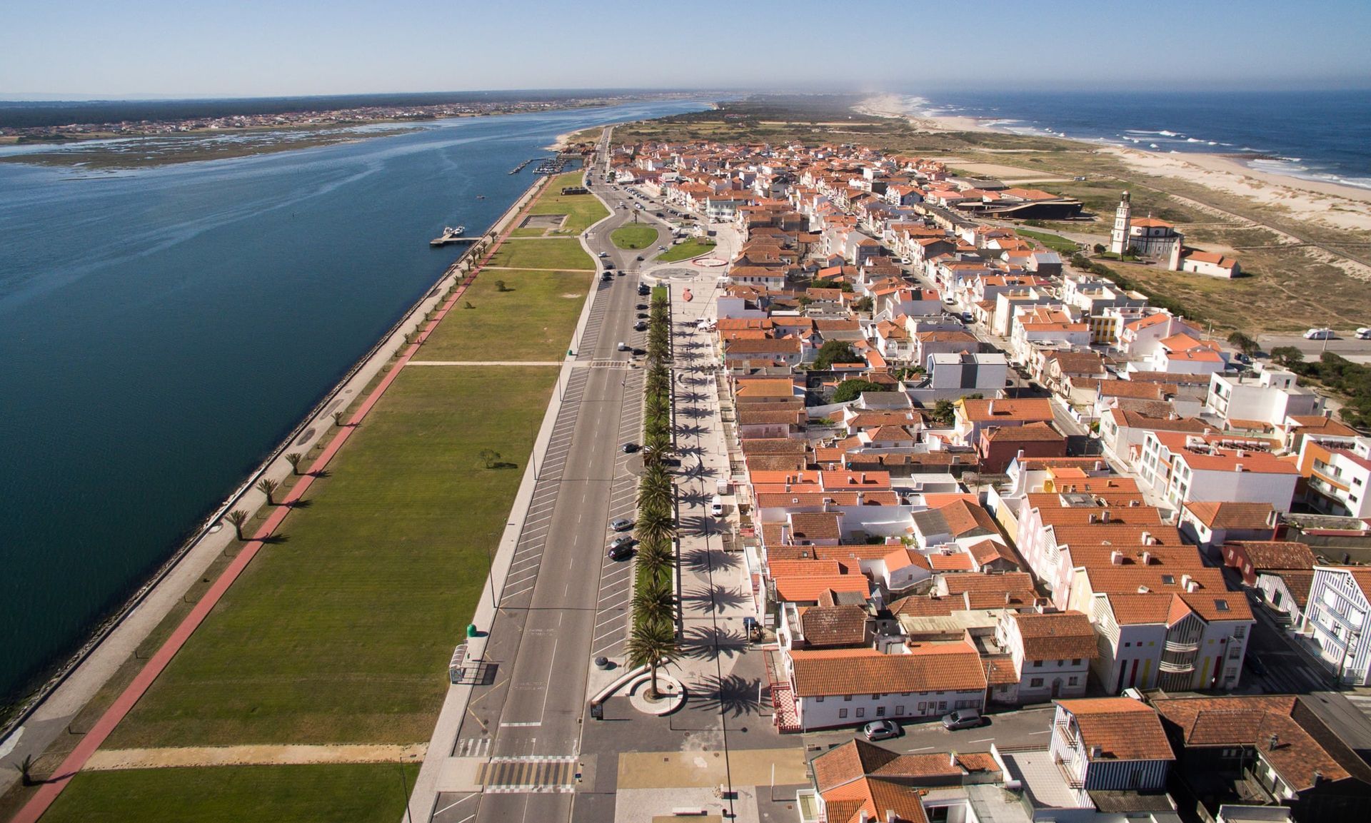 Aerial view of Costa Nova, featuring the Aveiro Lagoon on the left, the main palm-lined avenue and orange-roofed houses on the right, with the Atlantic Ocean in the distance.