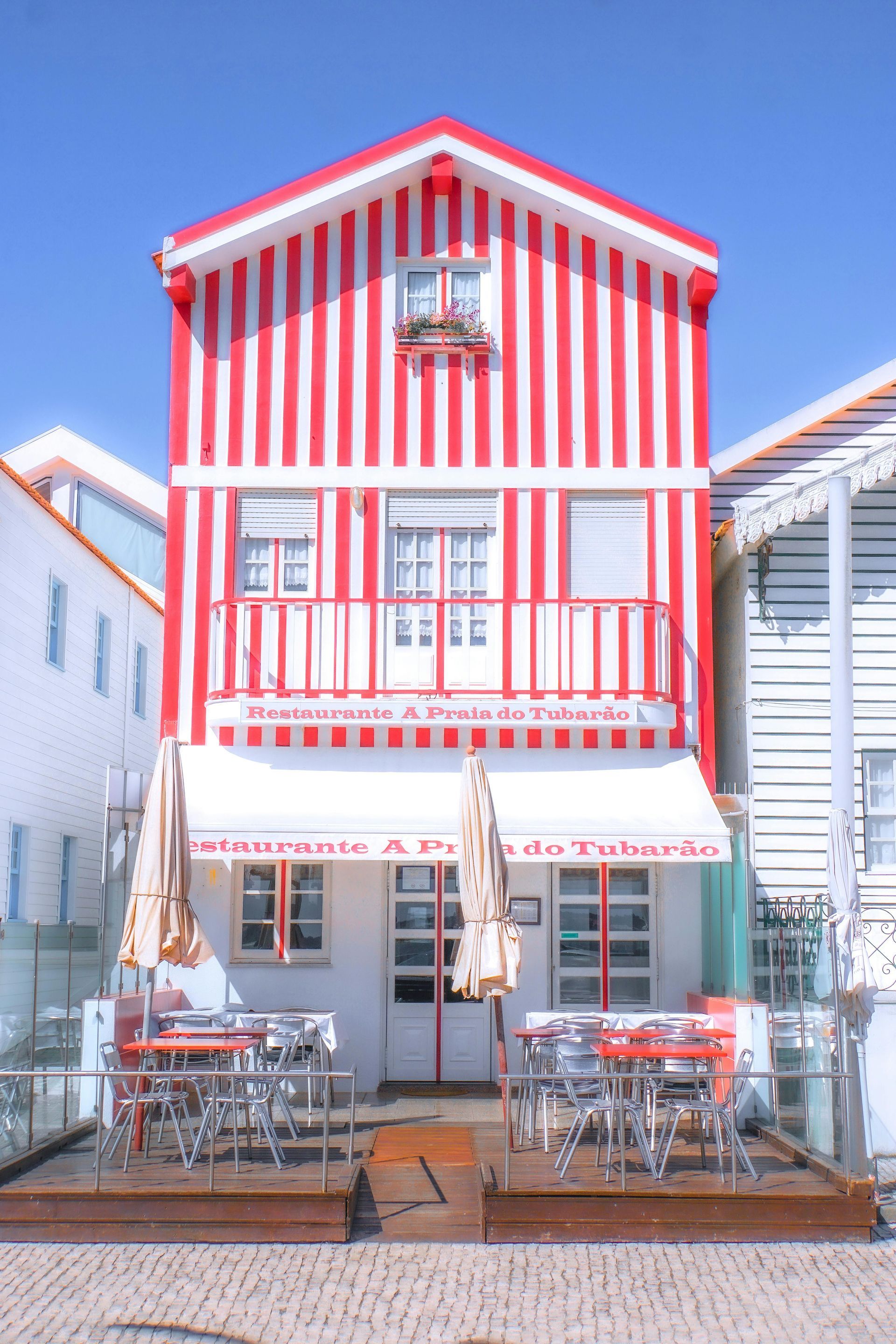 Traditional Costa Nova house with a red and white striped façade, clear blue sky in the background, and a terrace with tables and chairs in front.