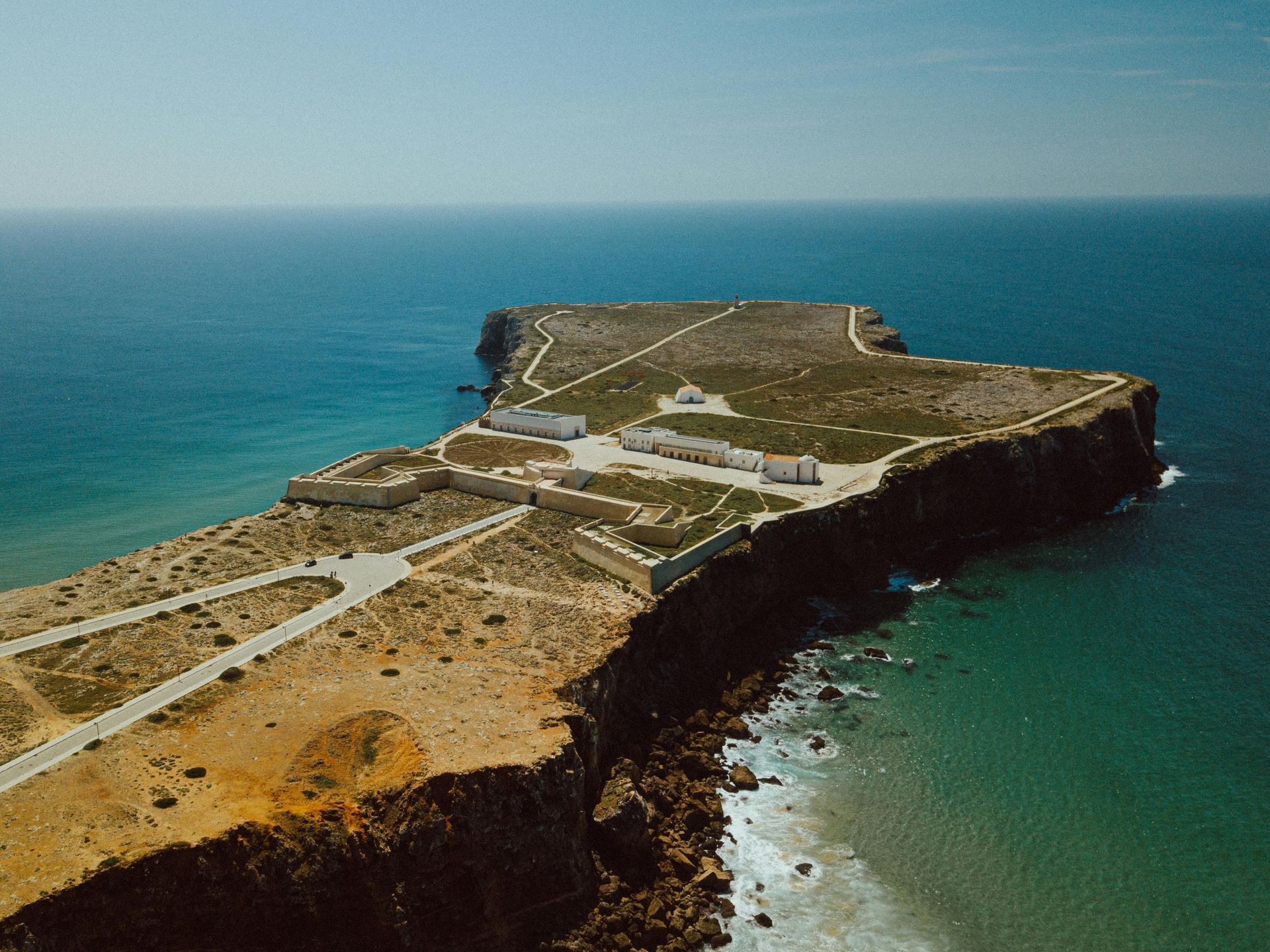 Wooden walkway over the sand at Costa Nova Beach, with the calm sea to the side and, in the distance, the Barra Lighthouse — one of the tallest in Europe — under a partly cloudy evening sky.