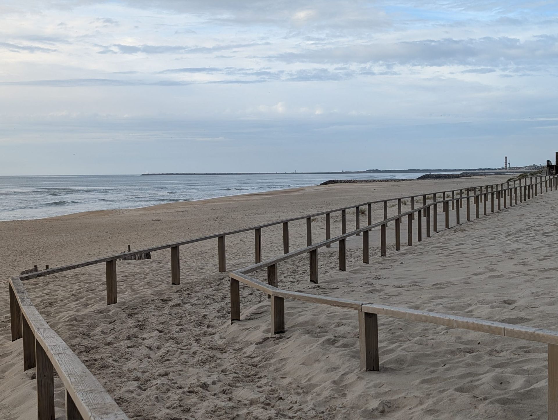Wooden walkway over the sand at Costa Nova Beach, with the calm sea to the side and, in the distance, the Barra Lighthouse — one of the tallest in Europe — under a partly cloudy evening sky.