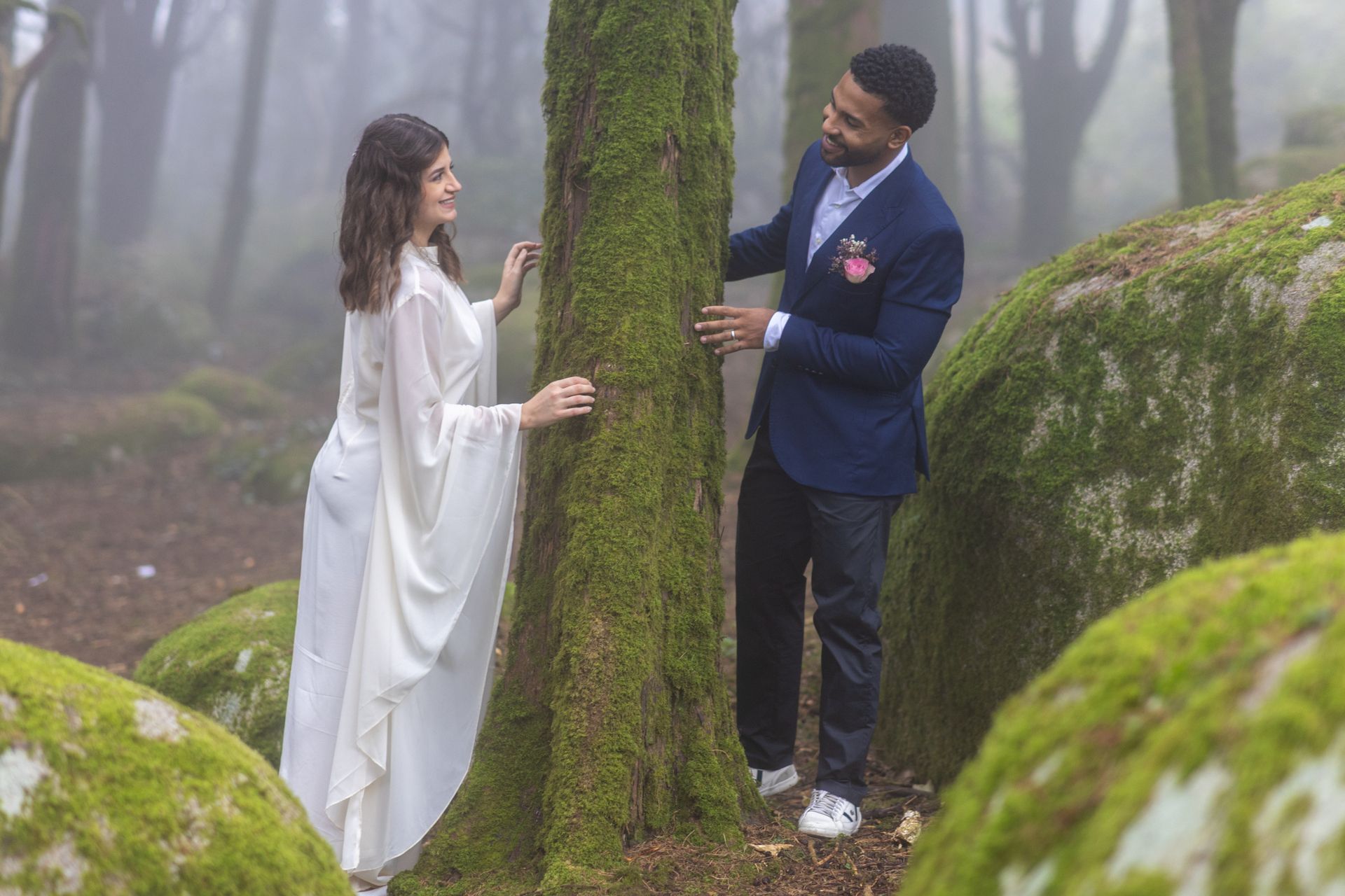 Casal romântico junto a uma árvore coberta de musgo, numa floresta mágica, envolta em nevoeiro. A noiva usa um vestido branco fluido e o noivo veste fato azul com flor na lapela.
