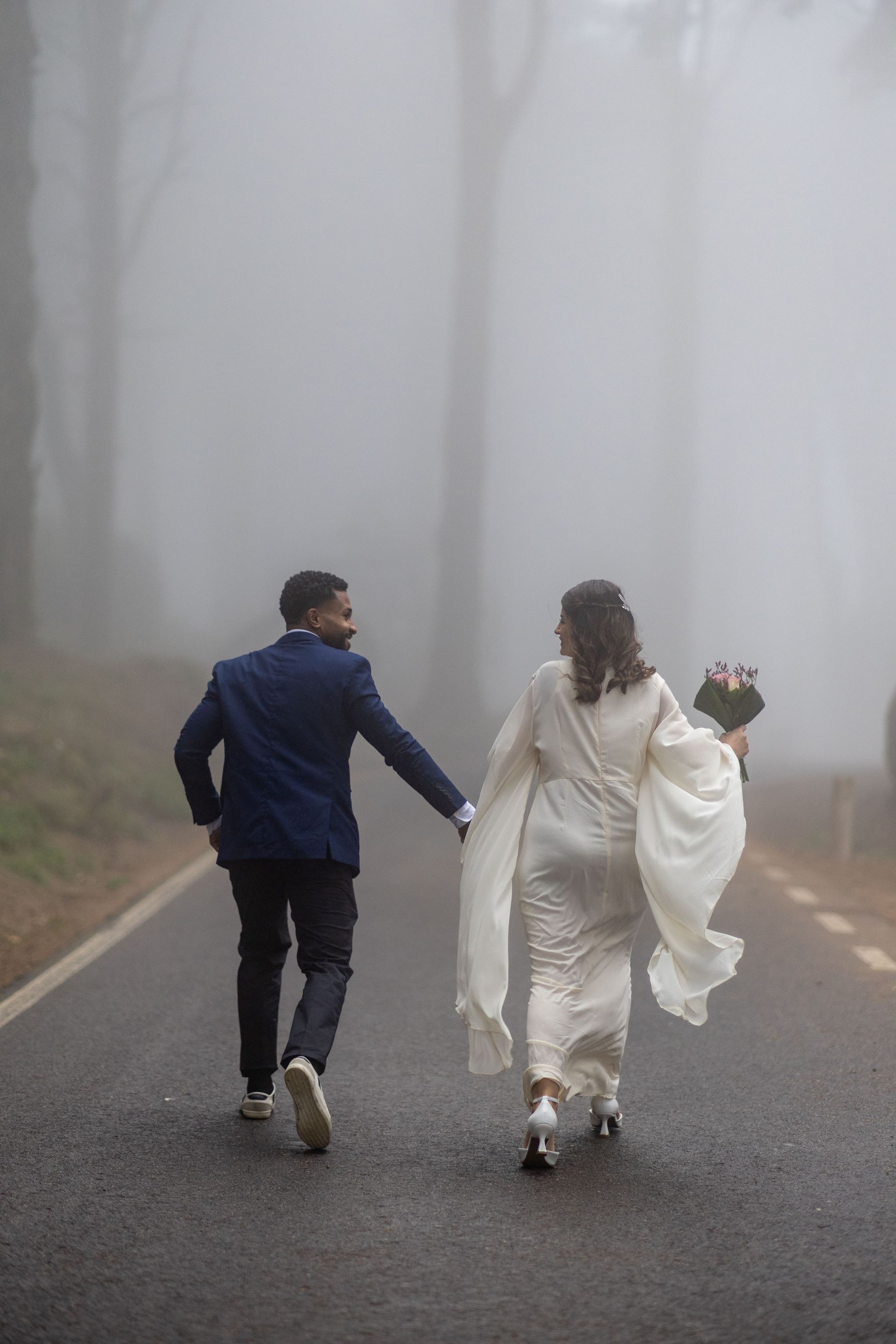 A couple, the bride holding a small bouquet, walking away holding hands down a foggy, tree-lined asphalt road. The low visibility adds a mysterious and romantic atmosphere. Peninha, Sintra.