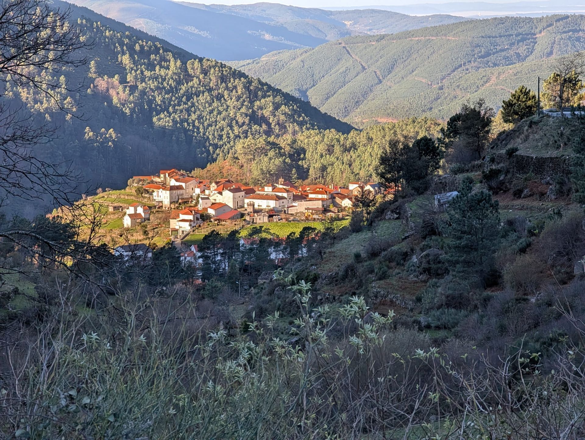 A scenic mountain village, Loriga, nestled in the Serra da Estrela region of Portugal. The village is made up of white houses with red-tiled roofs, clustered on a sunlit hillside surrounded by forested mountains and deep green valleys. The background reveals layered ridges of the mountainous terrain under a clear sky, highlighting the natural beauty of this alpine landscape.