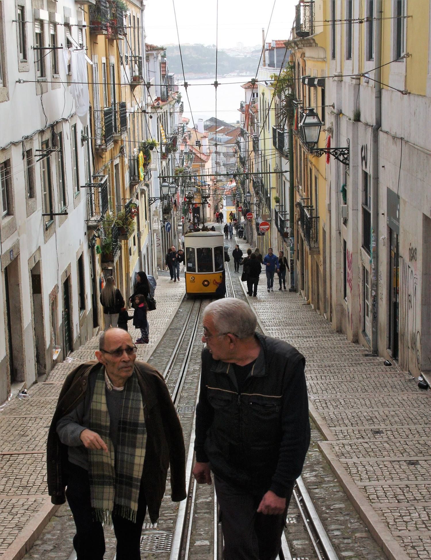 Two elderly men walking up Bica street in Lisbon, with the yellow tram and river in the background.