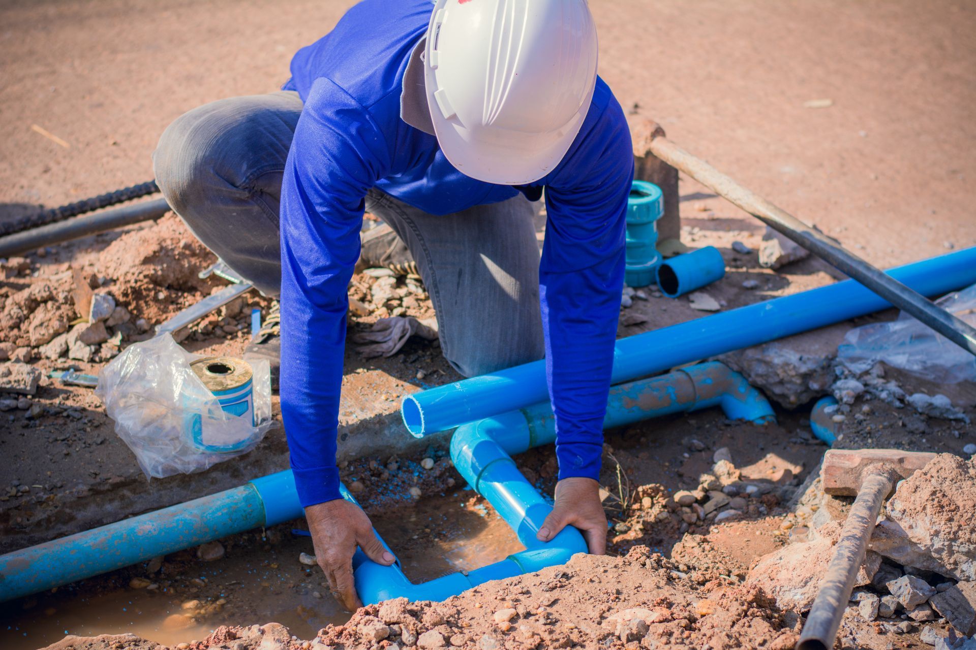 Plumber in blue shirt and white helmet connecting blue pipes in a dirt trench outdoors.