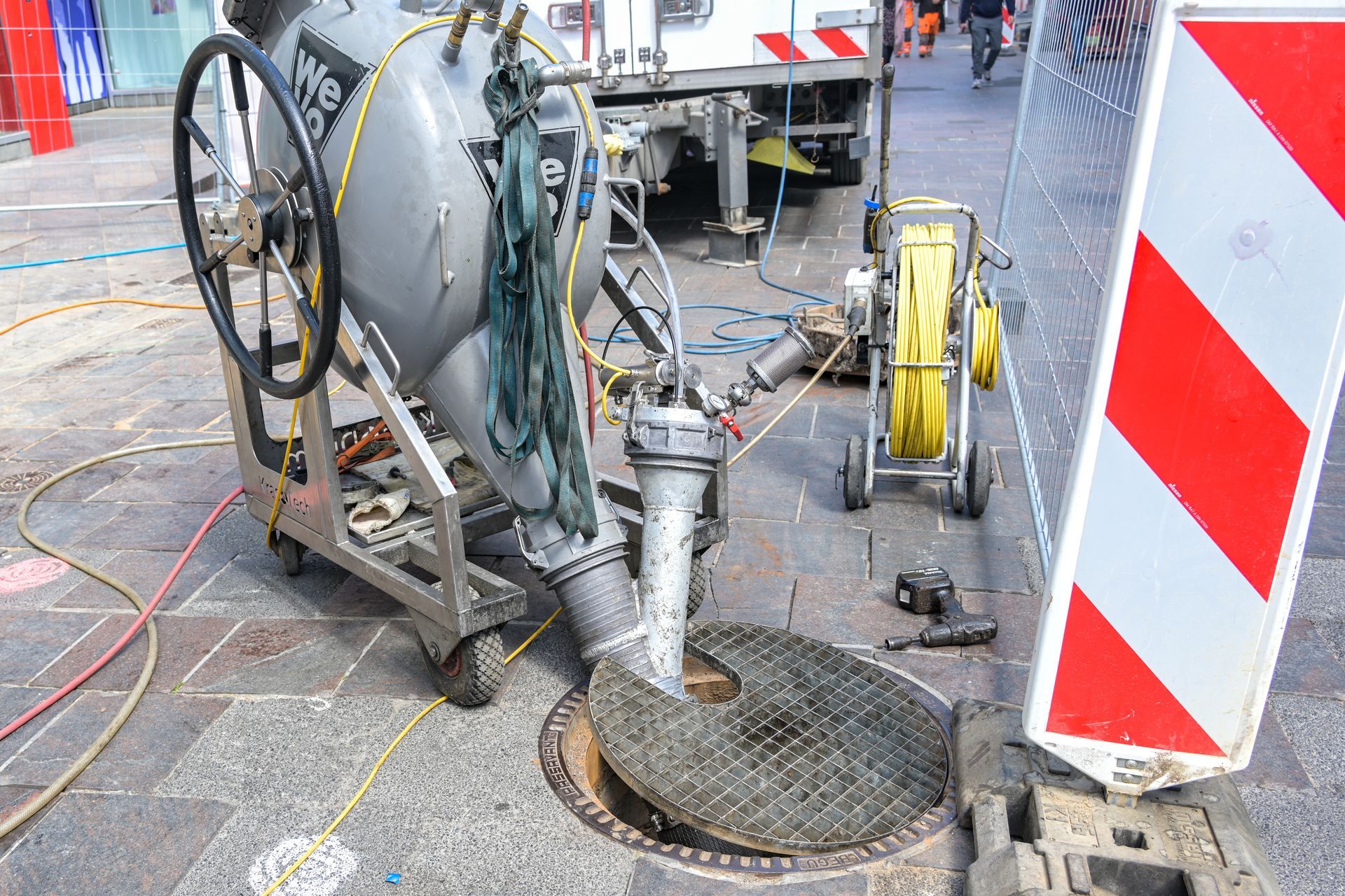 Equipment working on an open manhole in a city street. Grey machine, yellow hose, and safety barrier.