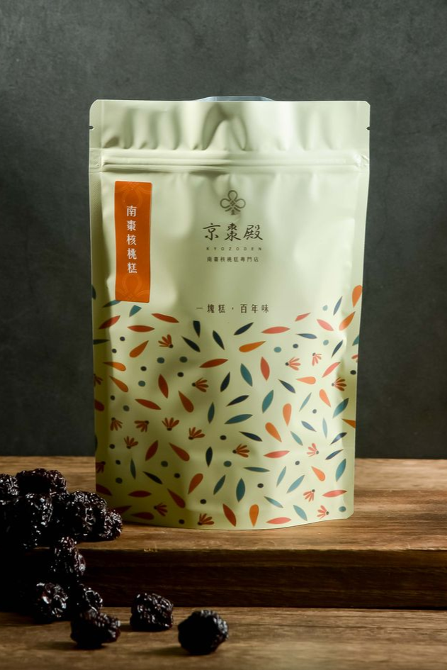 一個奶油色的立式茶包，上面印有色彩繽紛的葉子圖案，放在木質表面上，旁邊放著一些乾果。