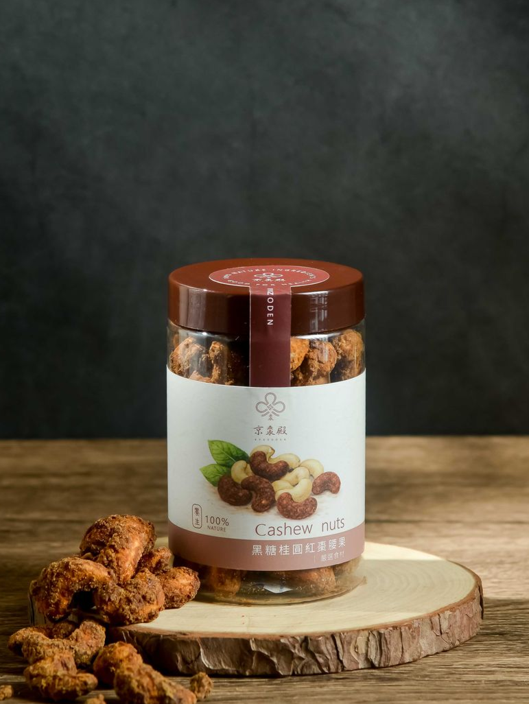 一罐透明塑膠罐裝的烤腰果放在一塊木板上，旁邊還放著一小堆散落的腰果。