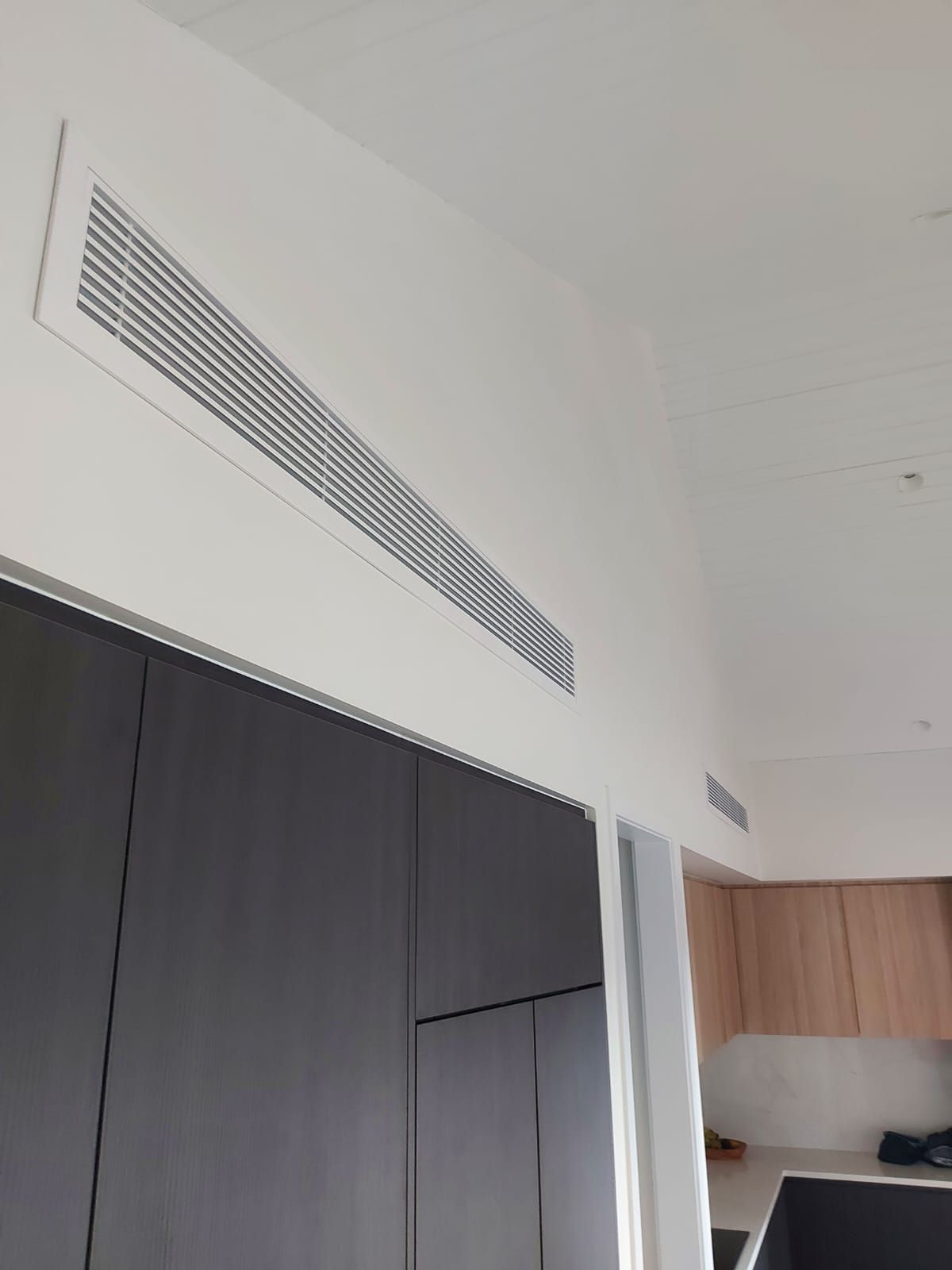 White rectangular air vent in the ceiling above a dark cabinet. Light wood cabinetry visible in the background.
