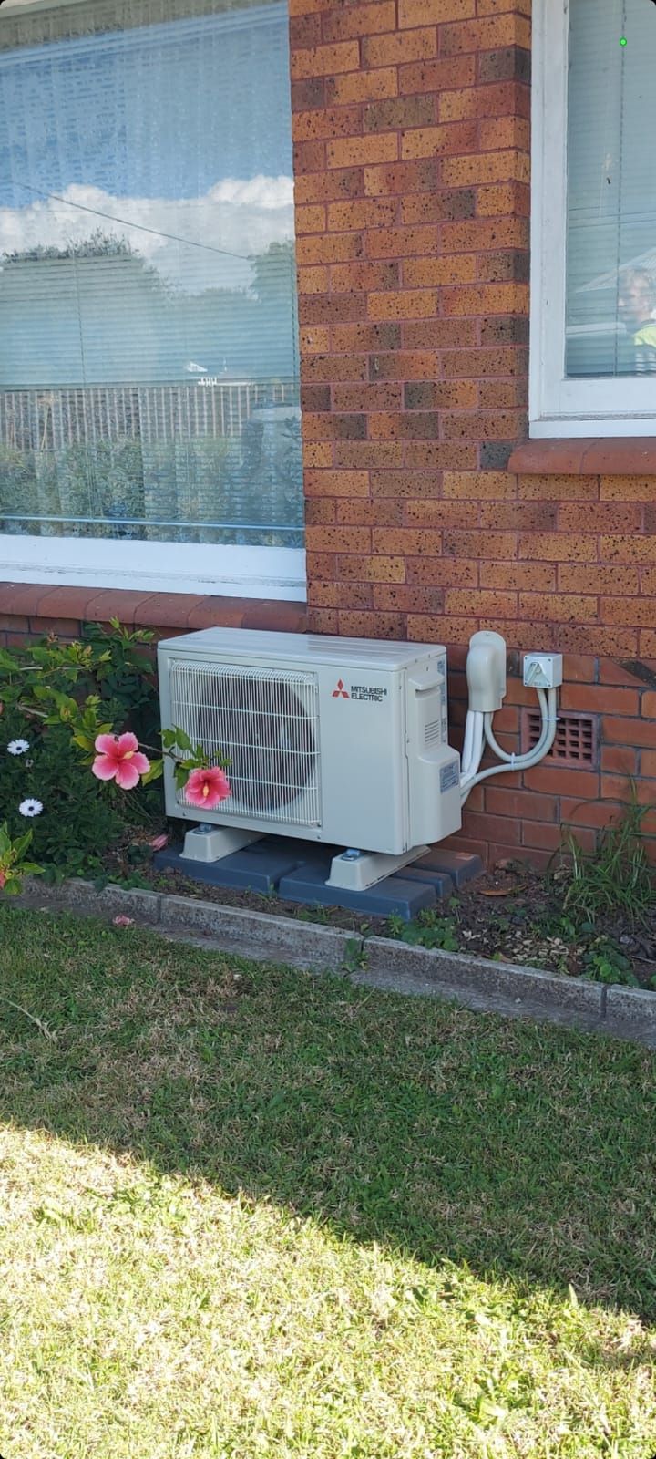 An air conditioning unit sits on a brick building's exterior, with green grass and a flower in the foreground.
