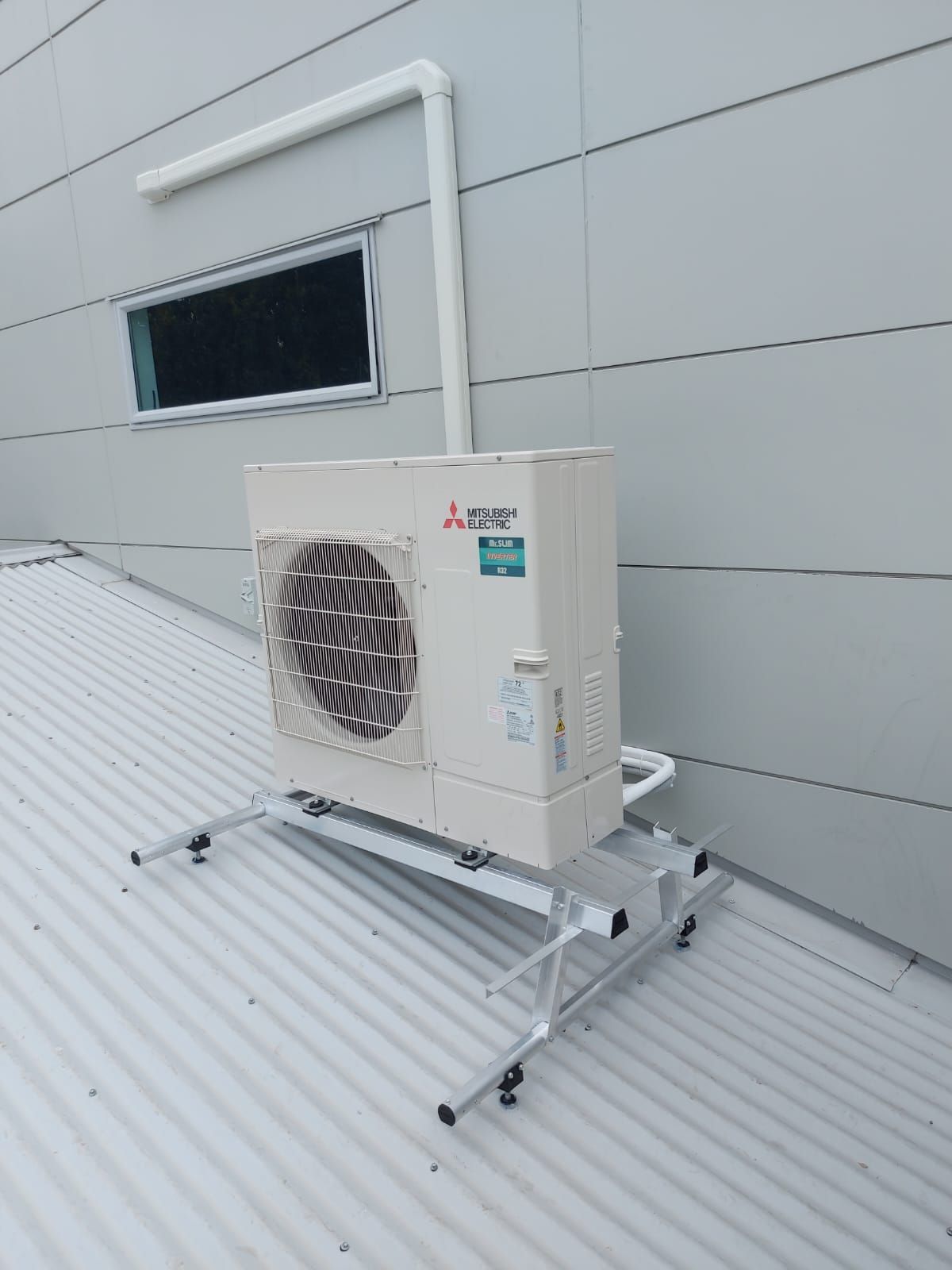 An air conditioning unit on a metal stand, mounted on a corrugated roof, with a white pipe running up the wall.