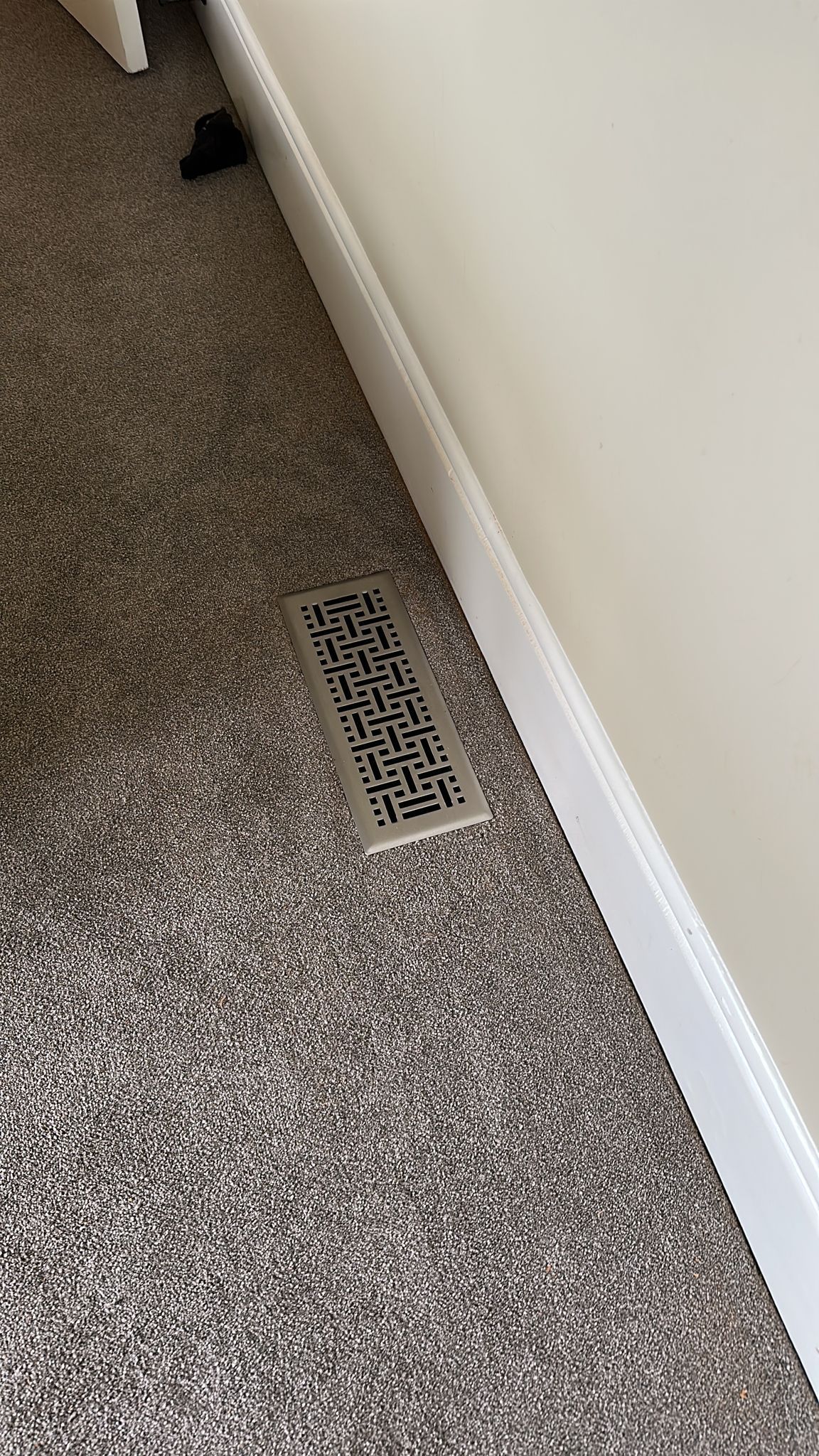 Metal floor vent in a room with gray carpet and a white wall.