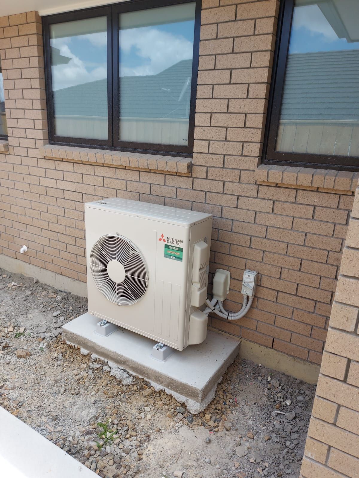Air conditioning unit mounted on a concrete pad against a brick wall, next to a window.
