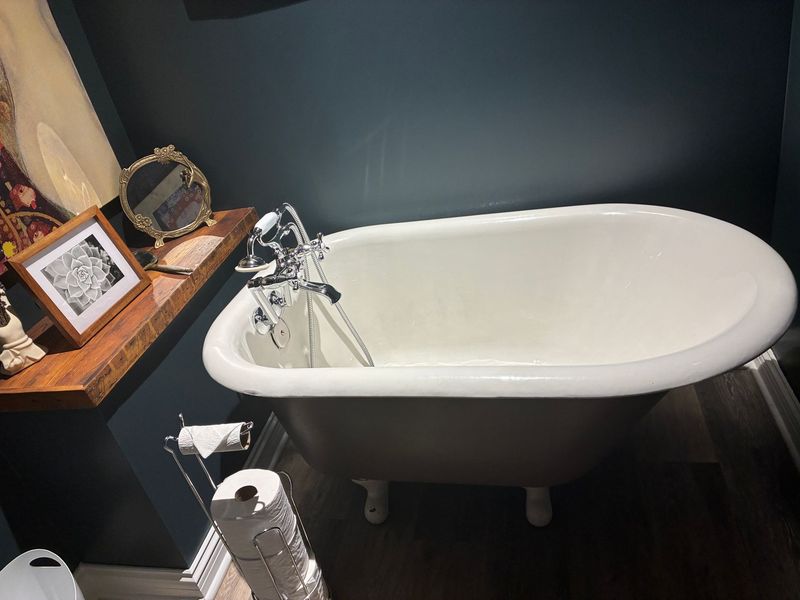 Clawfoot bathtub in a bathroom with a shelf holding a mirror and framed artwork. Dark teal walls.