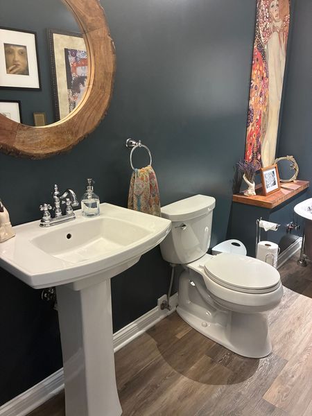A bathroom with a pedestal sink, toilet, dark teal walls, and a round mirror.