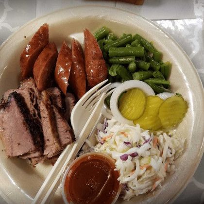Plate with sliced meat, sausages, green beans, coleslaw, pickles, and barbecue sauce on a table