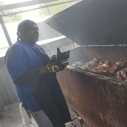 Person in blue shirt and black apron grilling meat over a large smoker outdoors.
