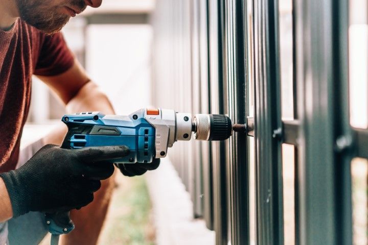 Person in black gloves using a blue and white power drill on a black metal fence outdoors.
