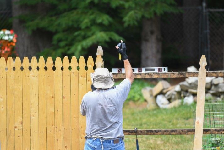 Man using level to build a wooden fence outdoors, holding a tool up, in front of greenery.