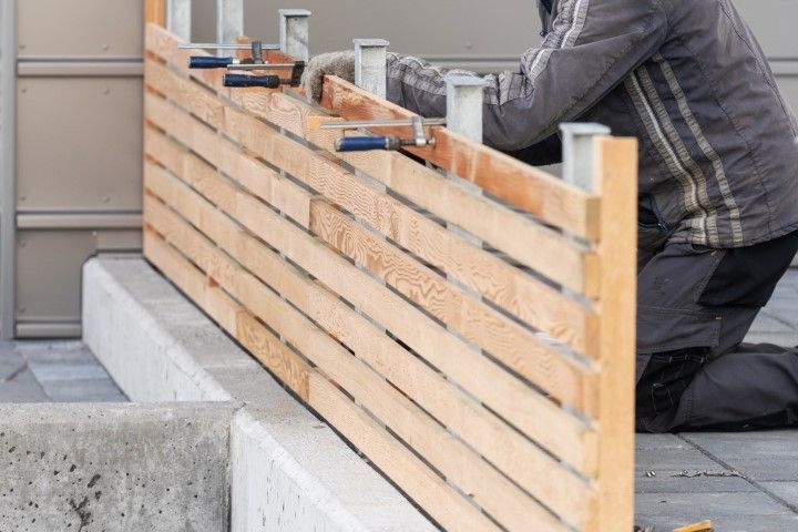 Person constructing a wooden fence with clamps, on a concrete base.