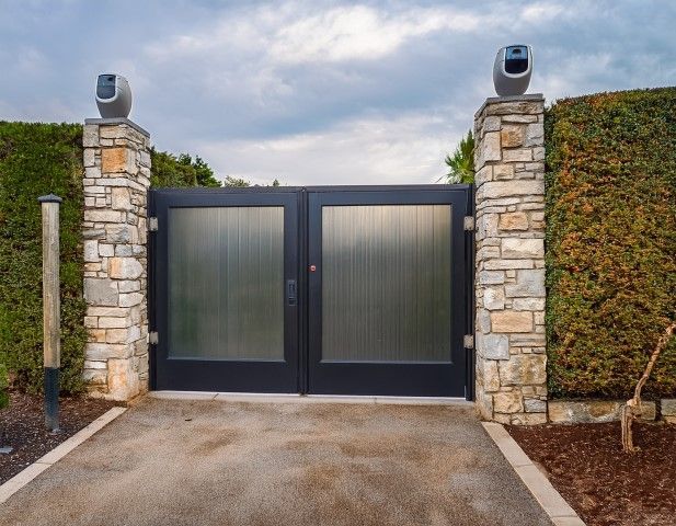 Stone pillars with automatic gate, concrete driveway, and green hedges.