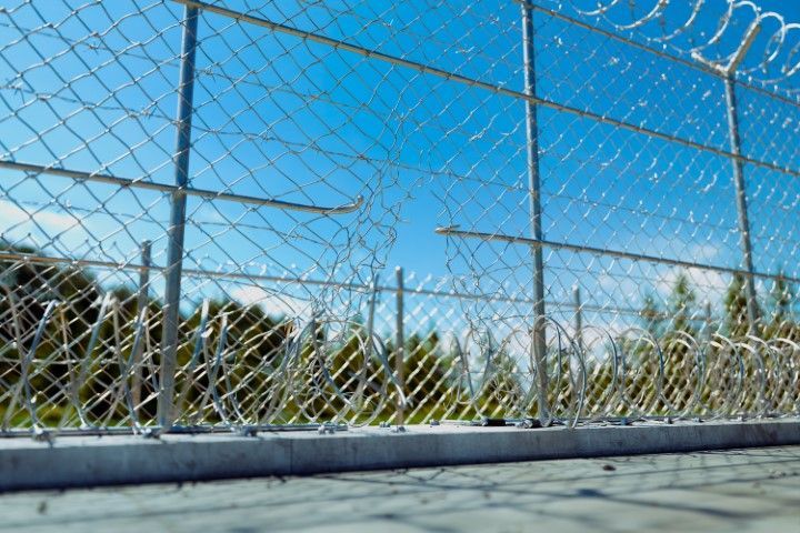 Chain-link fence with barbed wire against a blue sky, a hole visible in the mesh.