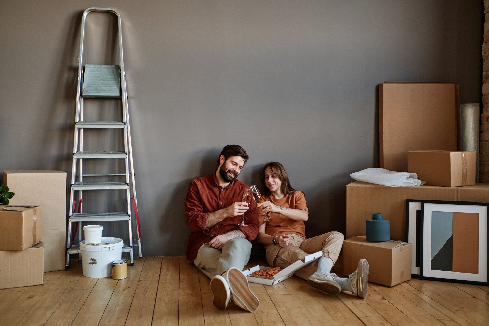A man and a woman are sitting on the floor eating pizza and drinking wine.