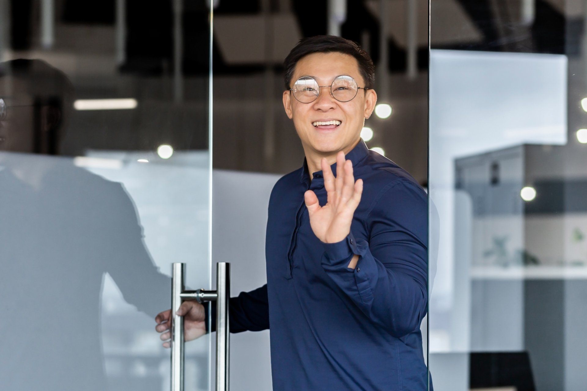 A man wearing glasses is waving from behind a glass door.