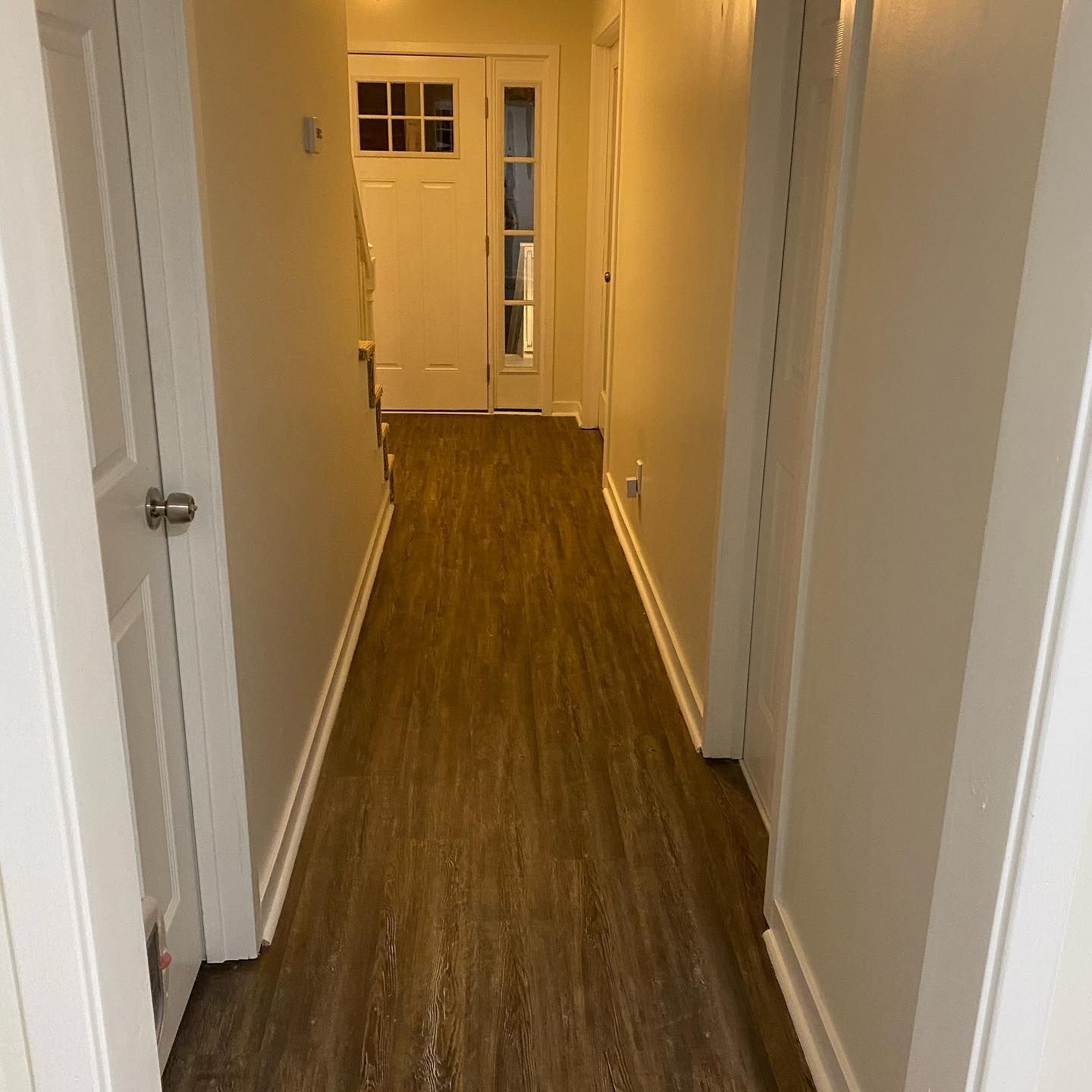 A long hallway with wooden floors and white walls leading to a door.