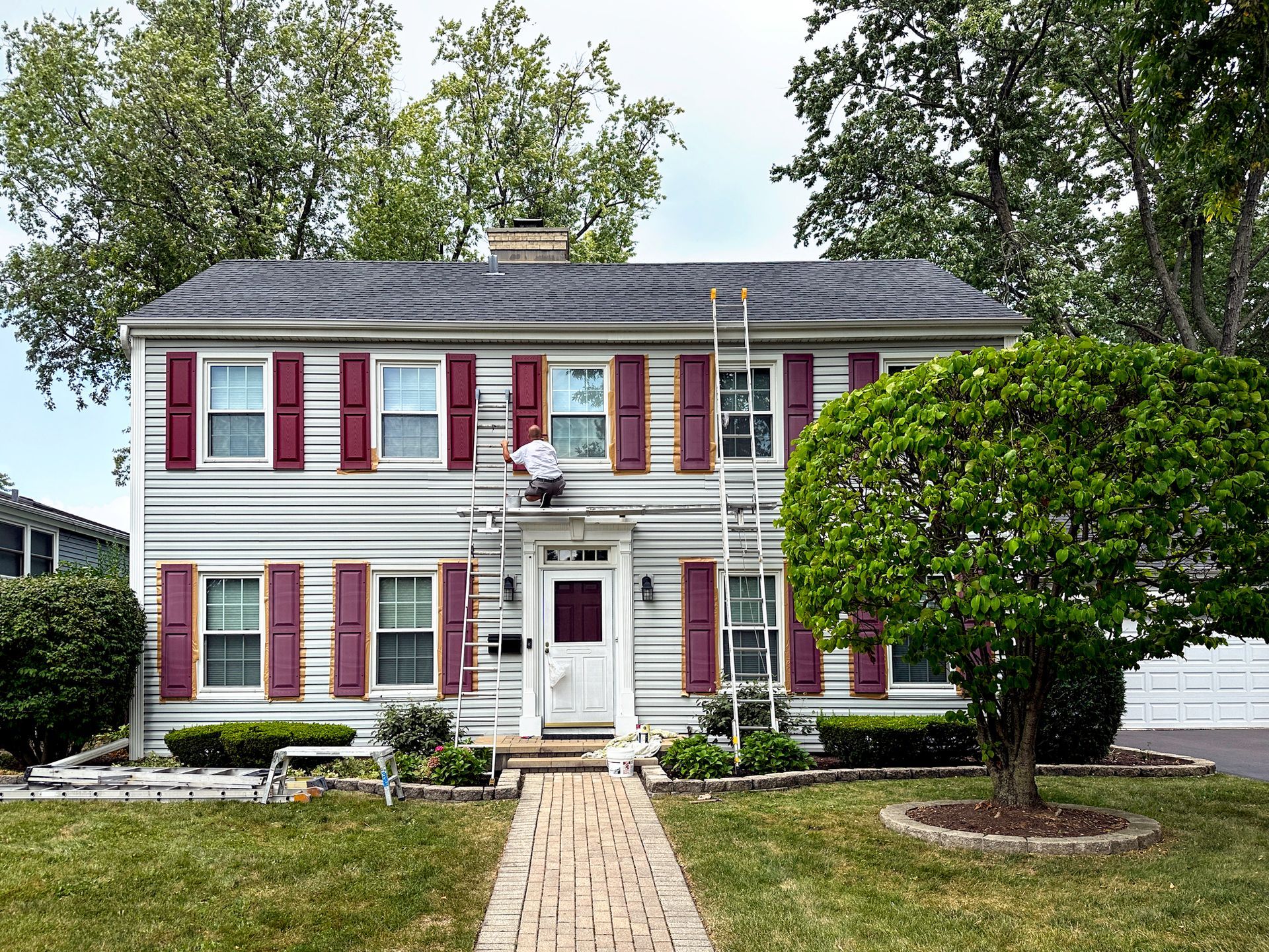 A man is painting the side of a house with red shutters.
