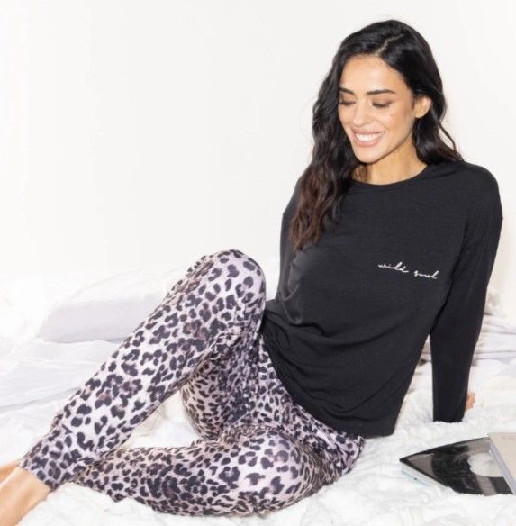 Una mujer con un top negro y pantalones con estampado de leopardo sonríe, sentada en la cama.
