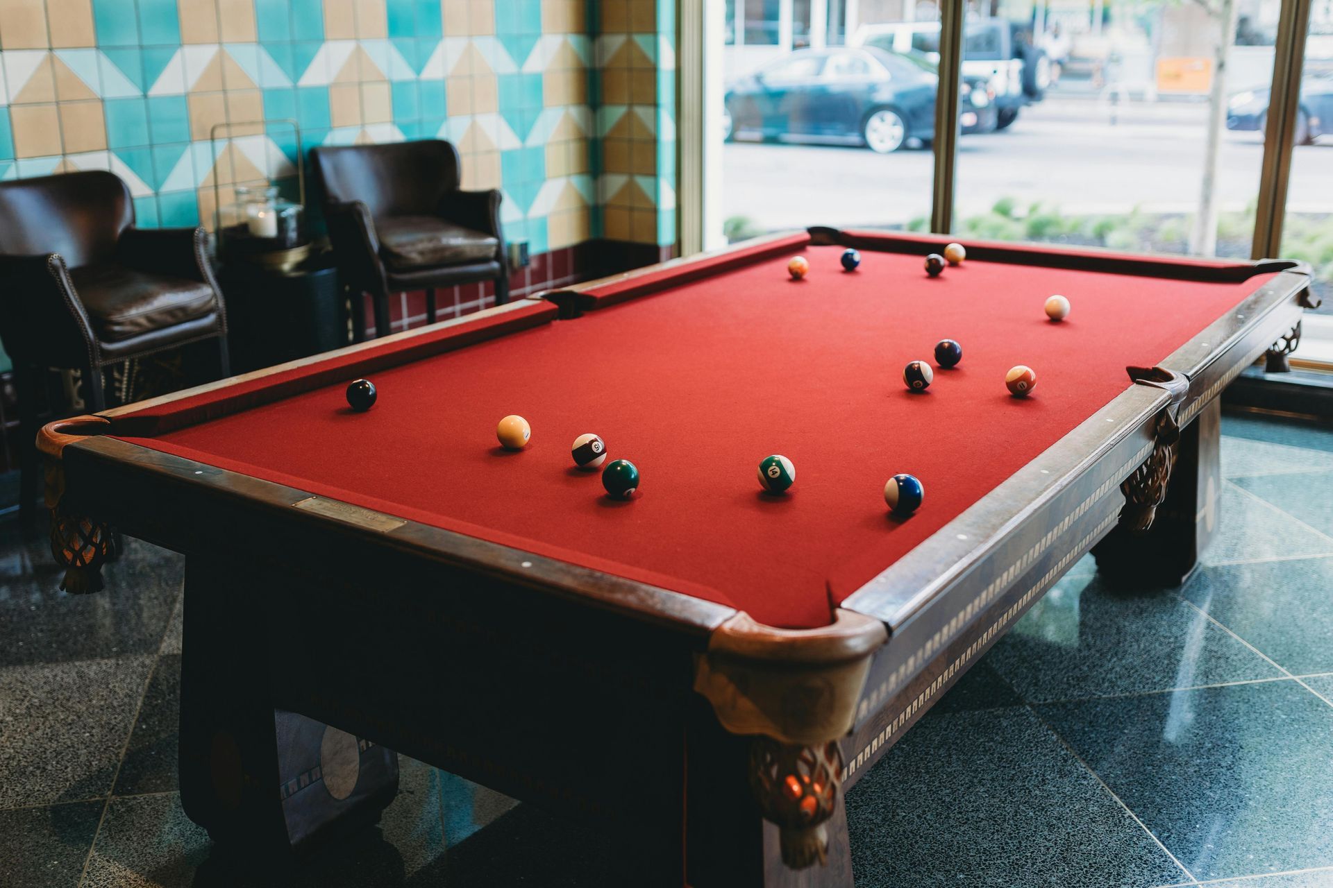 Pool table with red felt and scattered balls in a lounge by large windows