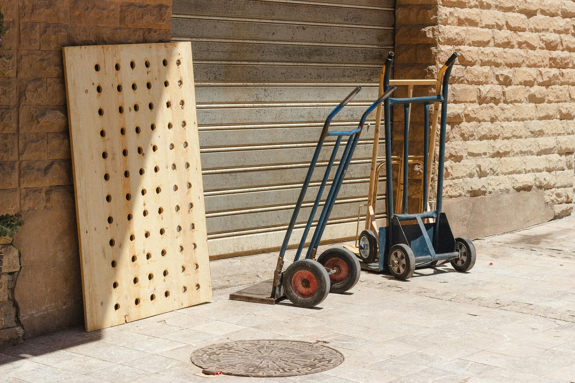 Two hand trucks and a perforated wooden board leaning against a brick wall in a sunny alley