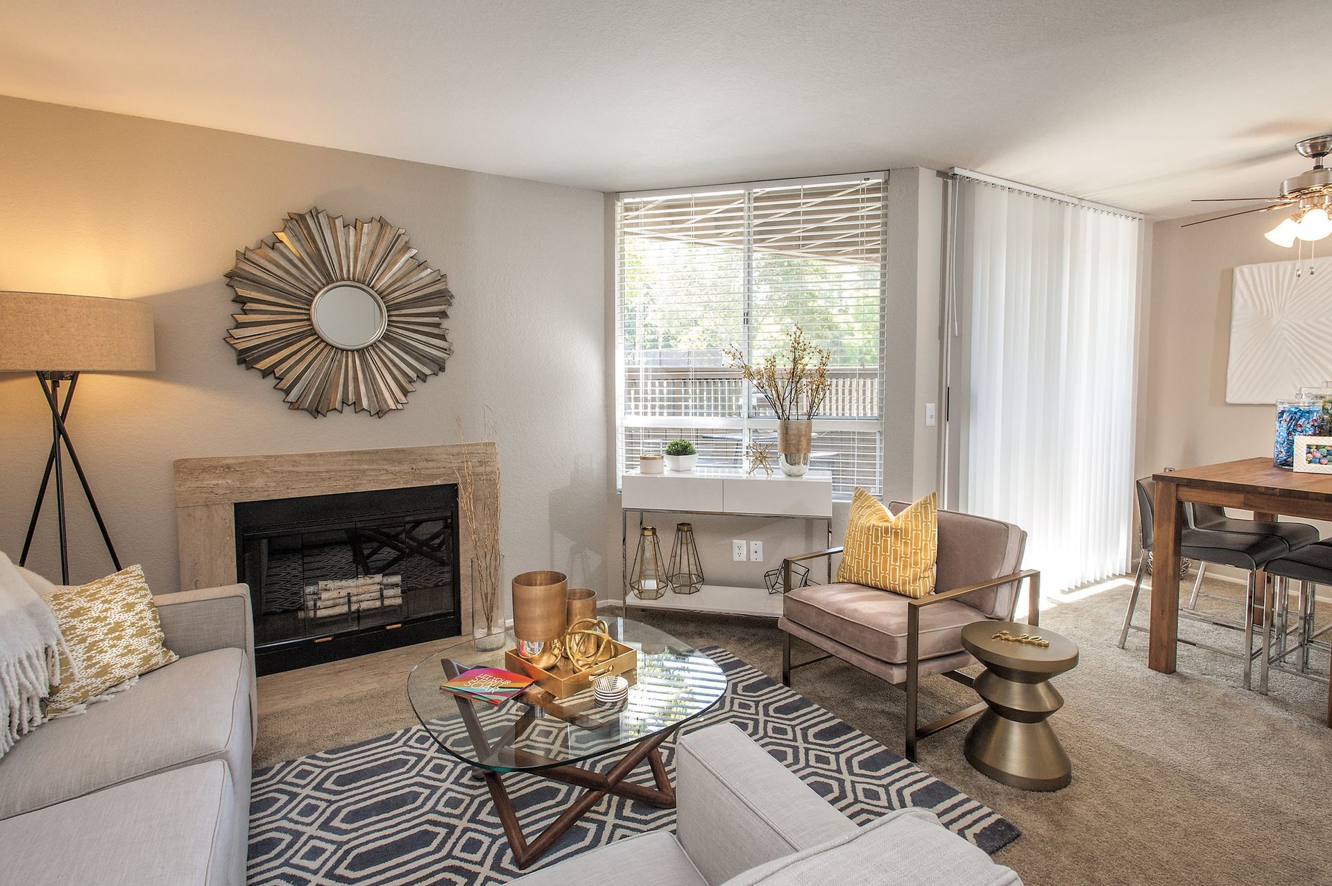 Living room in an apartment with a beige sofa, fireplace, glass coffee table, and a dining area by a large window.