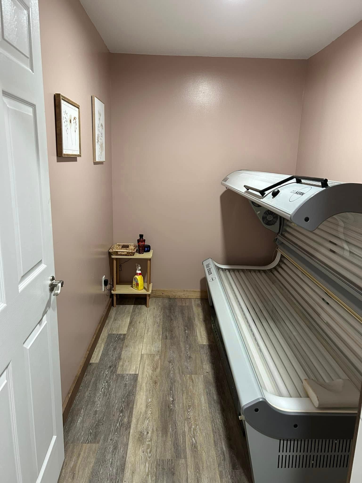 A tanning bed sits in a small room with light pink walls, wood-look flooring, and a small corner shelf with supplies.