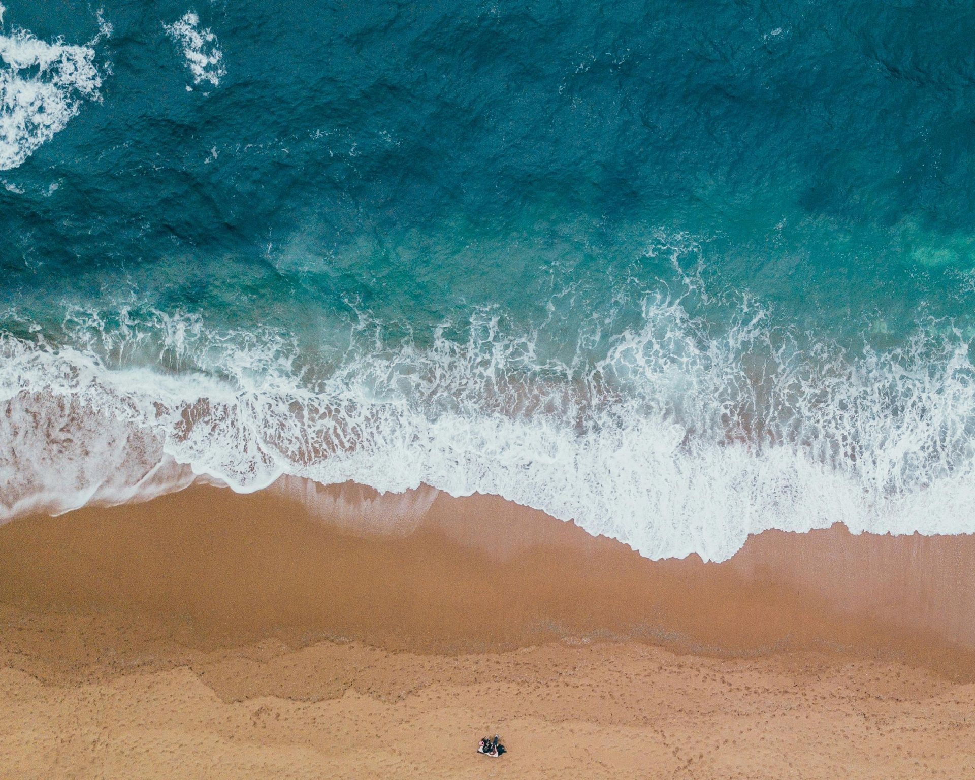 Aerial view of waves crashing against a sandy beach with a person walking along the shore.