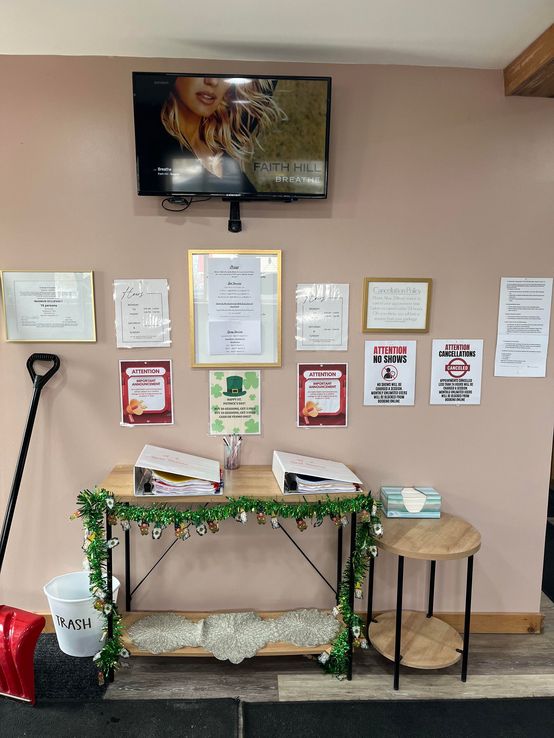 A wall-mounted TV over a small table holding two binders, decorated with a green garland and flyers on the wall behind.