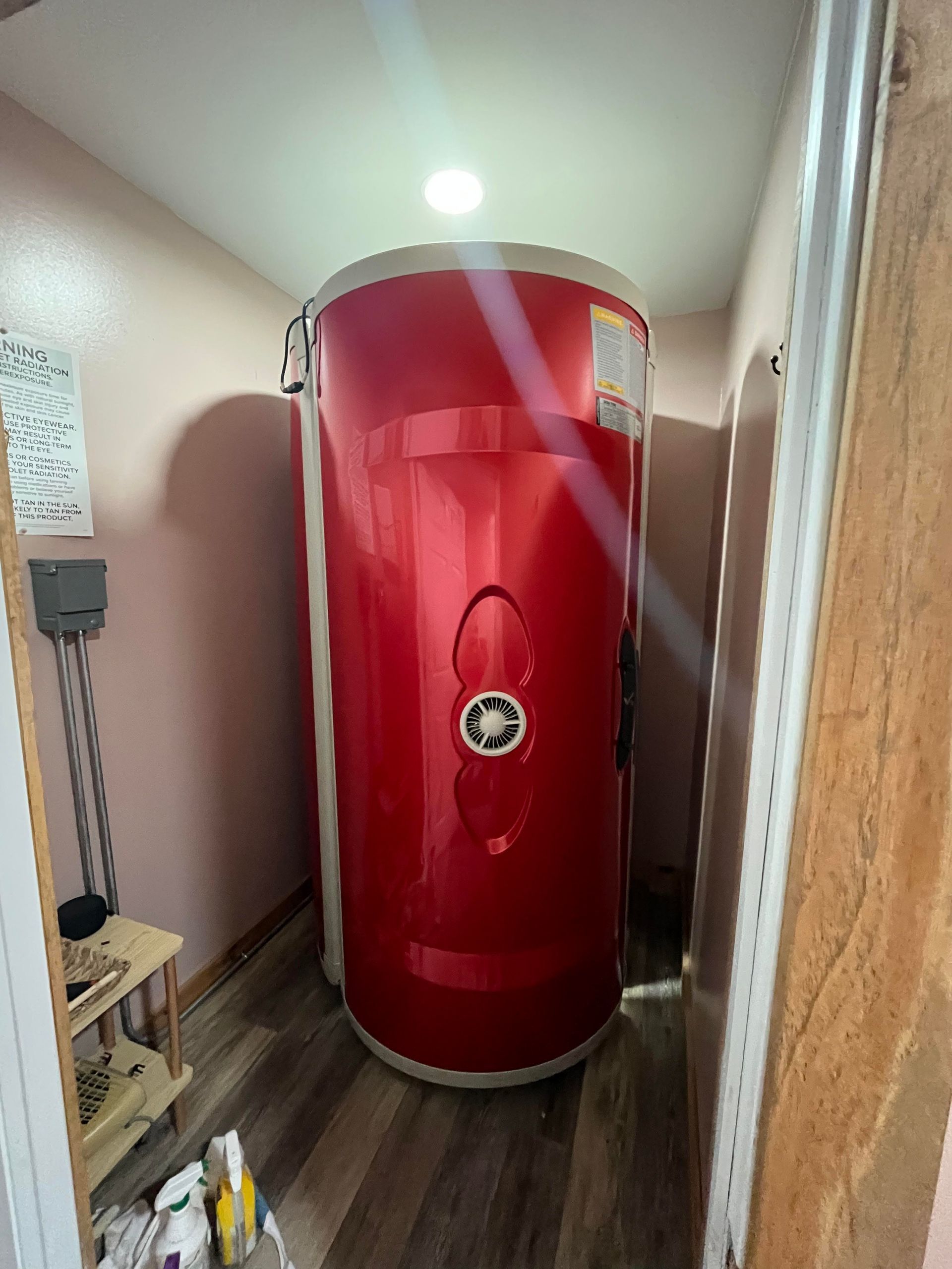A red, cylindrical stand-up tanning bed centered in a small, wood-floored room next to a small shelving unit.