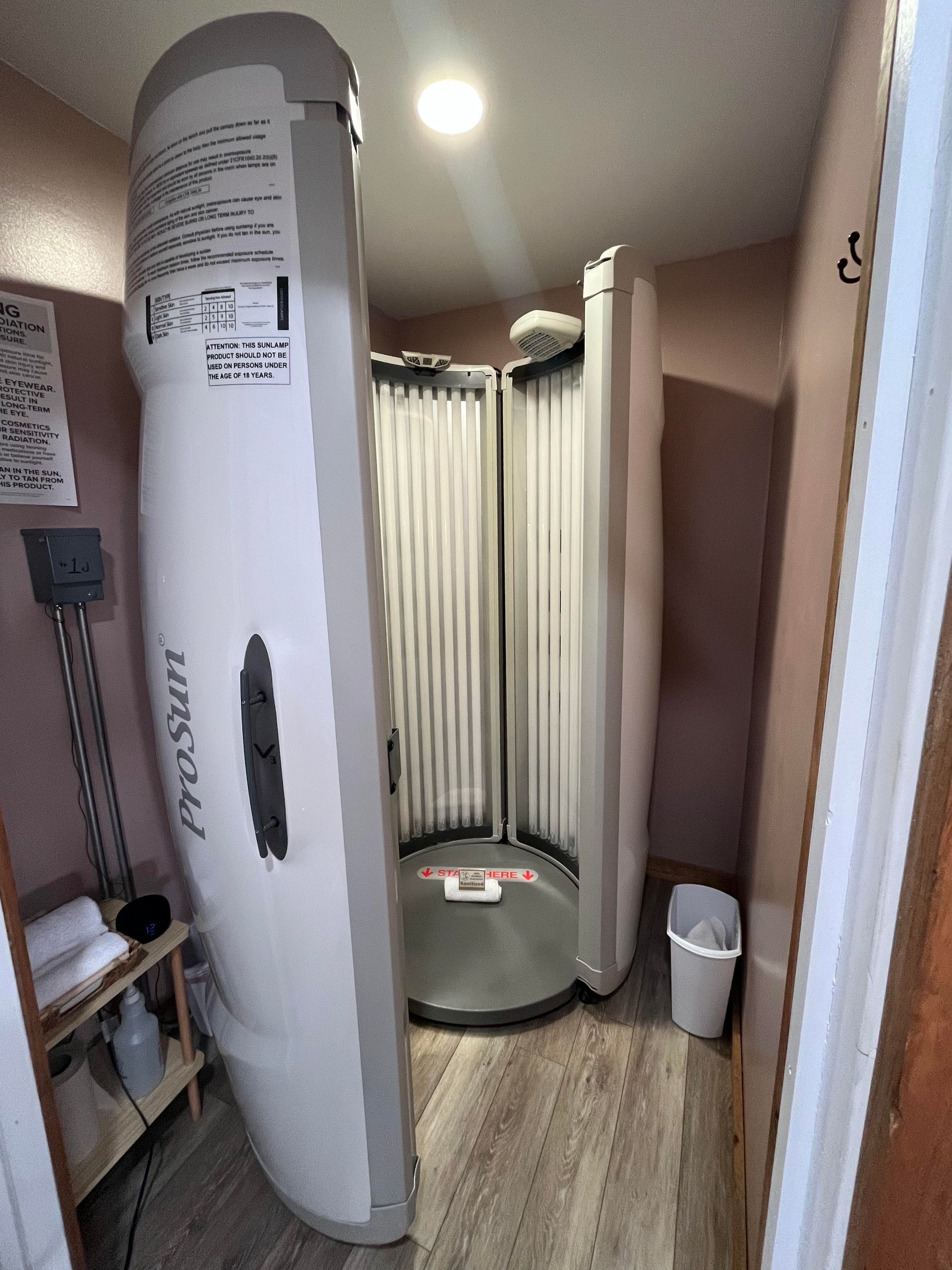 A stand-up ProSun tanning booth inside a small, neutral-toned room with wood-look flooring and a trash can.