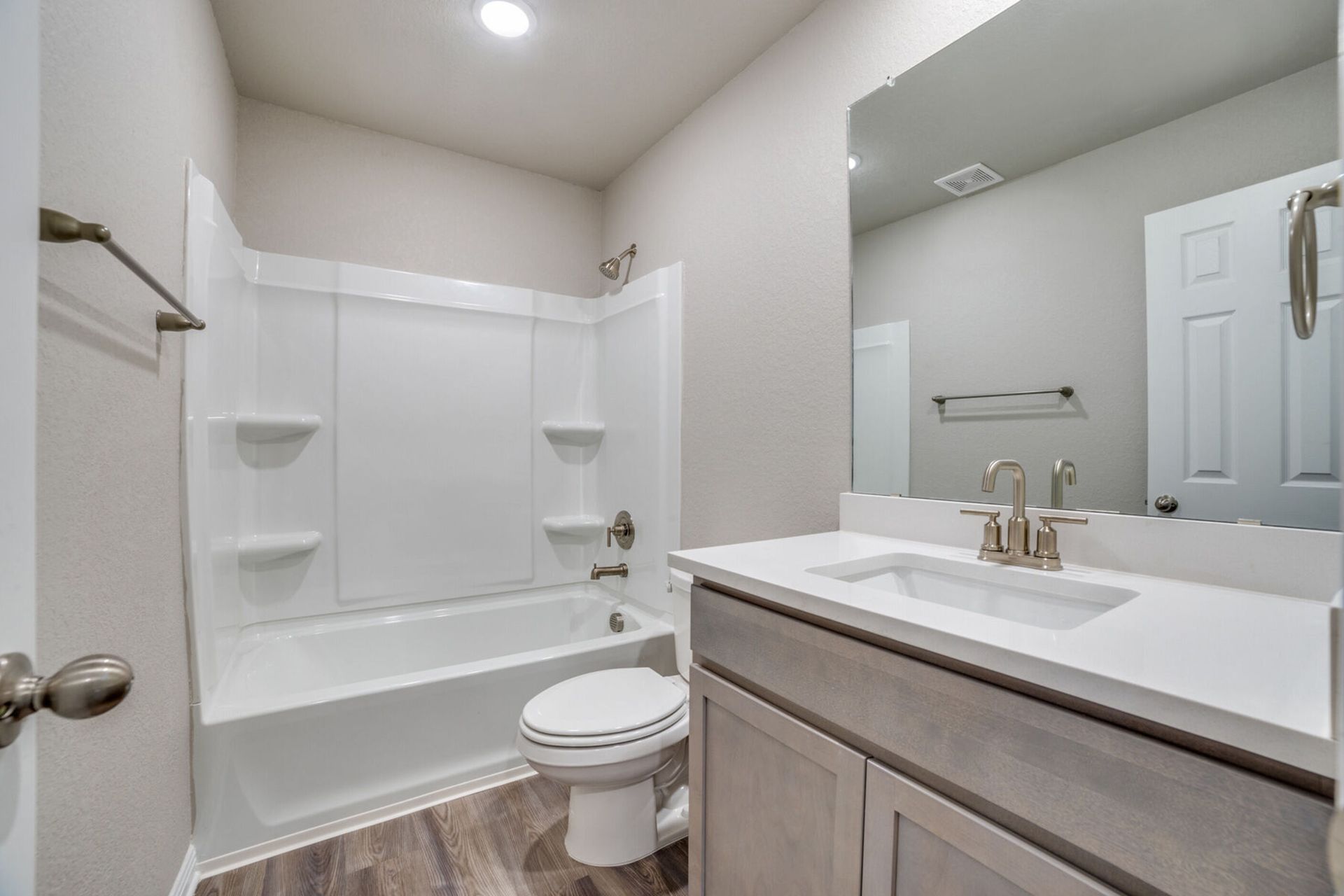 Bathroom with white tub/shower, toilet, vanity, and large mirror; light gray walls and wood-look floor.