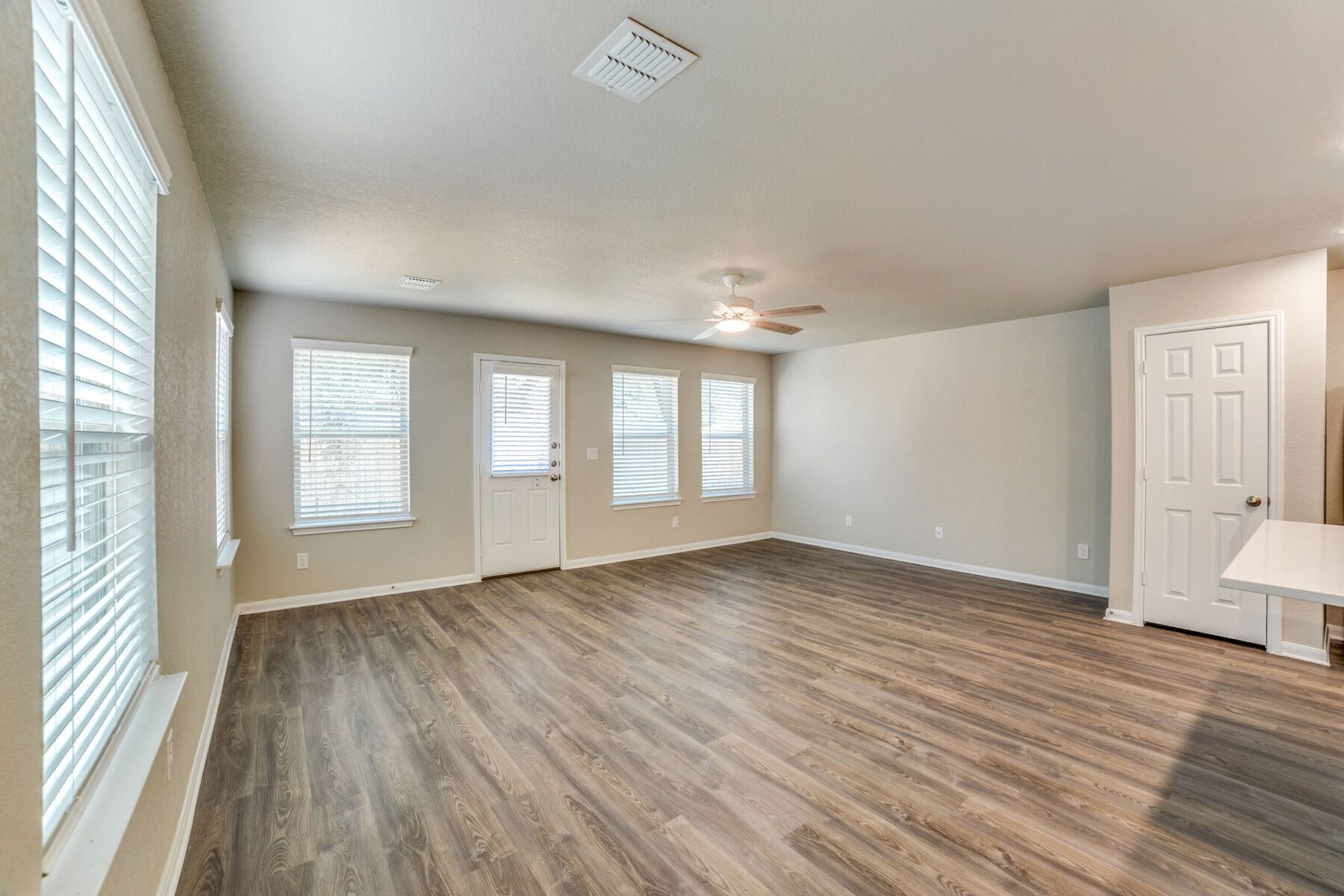 Empty living room with wood-look floors, beige walls, and windows with blinds.