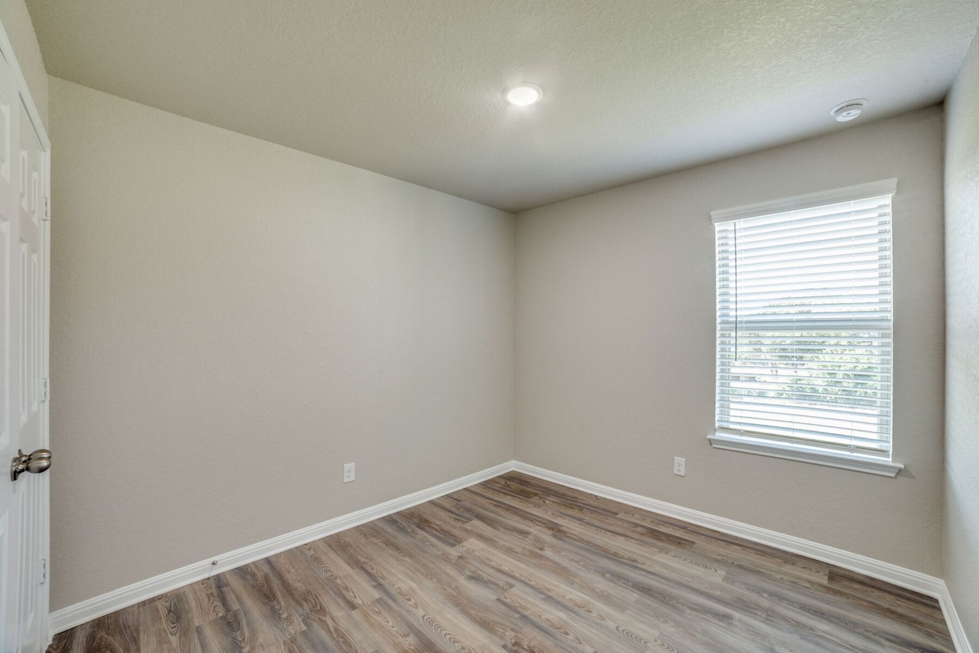 Empty room with light-colored walls, wood-look floor, window with blinds, and a closed door.