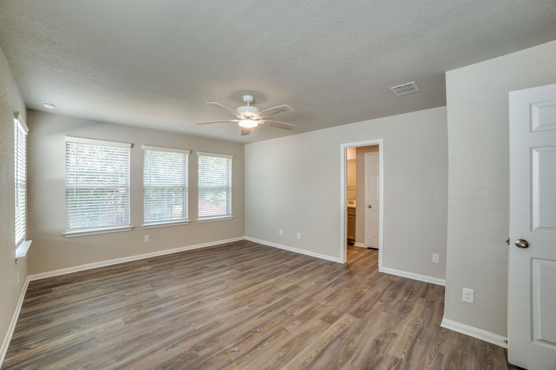 Spacious bedroom with gray walls, wood-look floors, and three windows; door to ensuite and white door to the right.