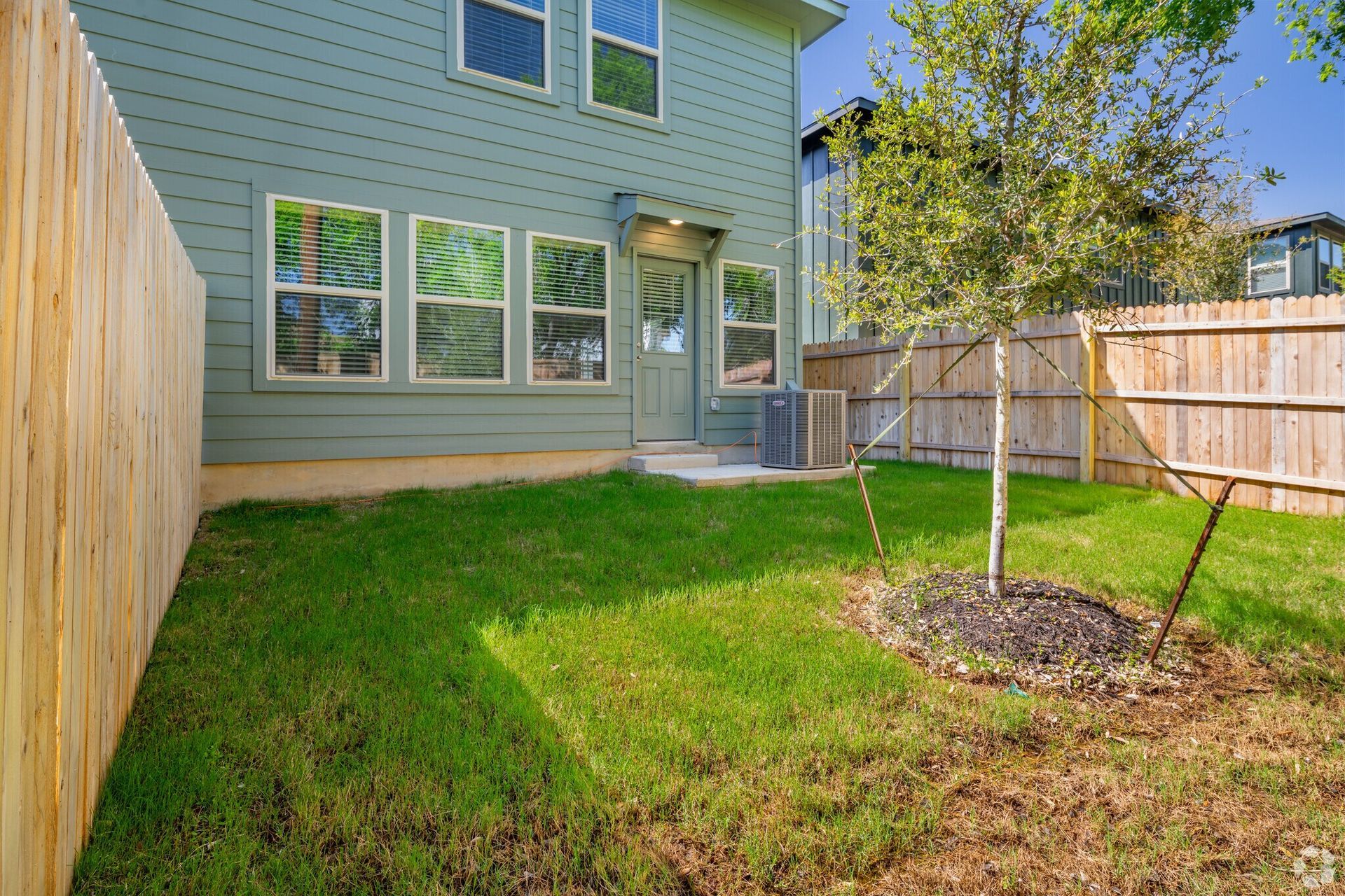 Backyard of a house with green grass, a tree, and a wooden fence.