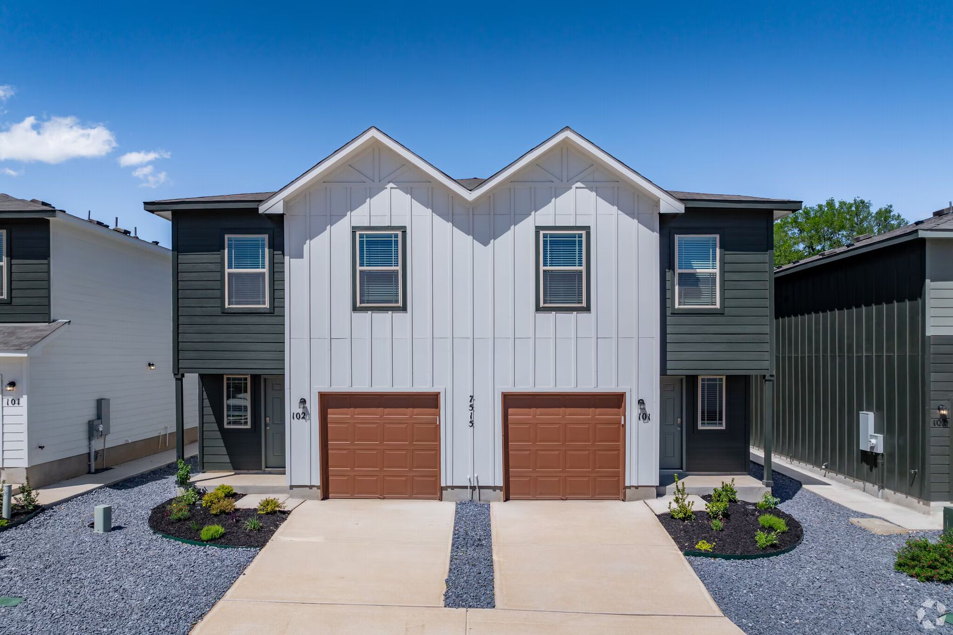 Two-story duplex with white and dark gray siding, brown garage doors, and blue sky.