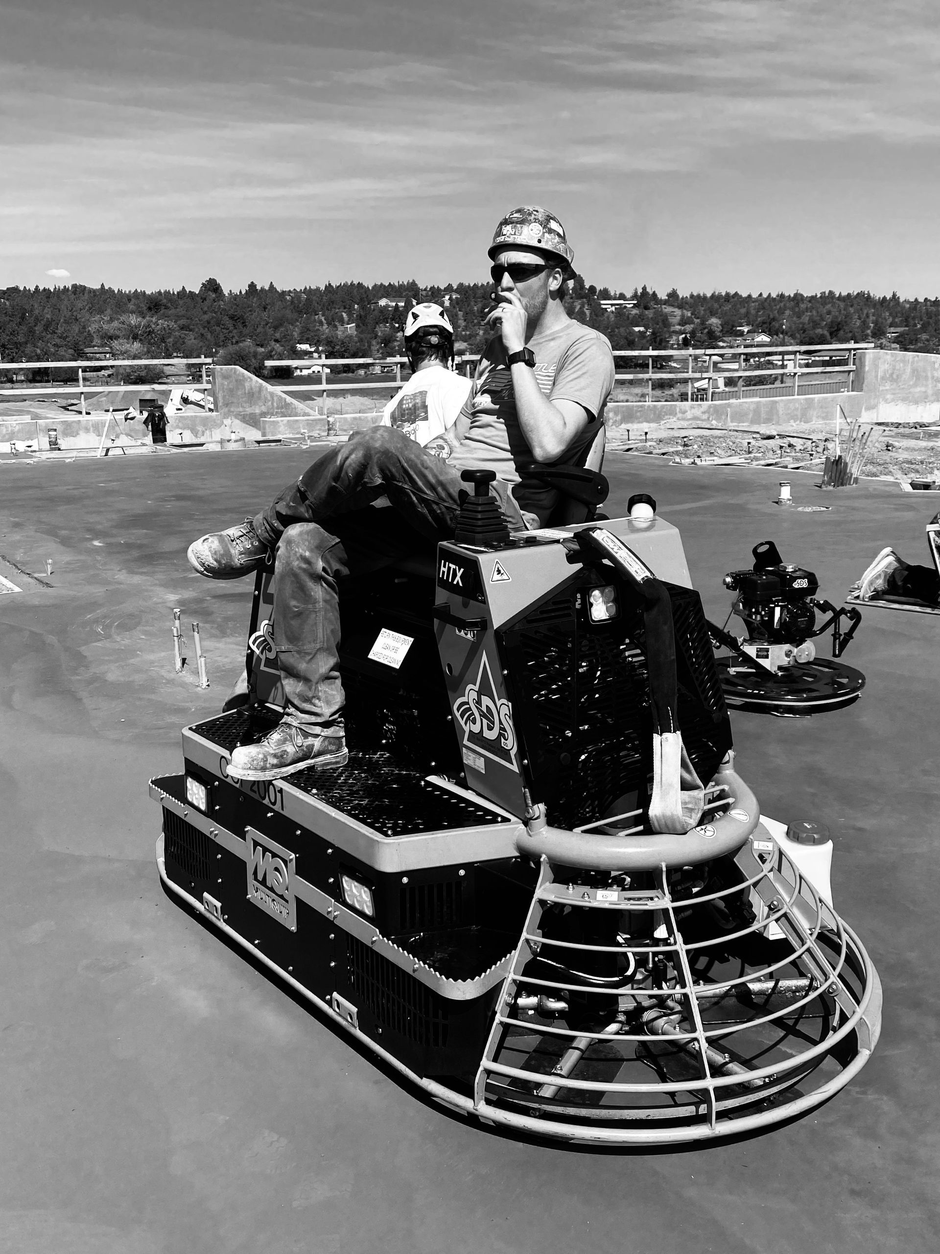 Two construction workers operating a power trowel on a rooftop construction site under a bright sky.