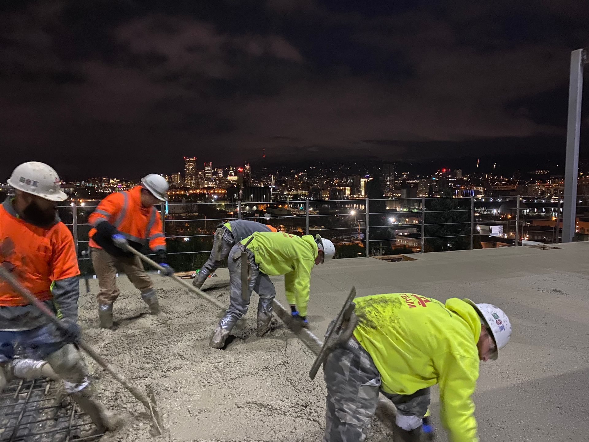 Construction workers leveling concrete at night with cityscape backdrop.