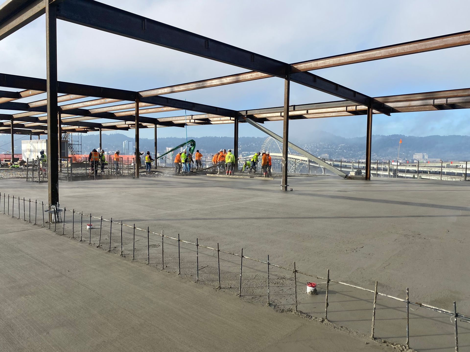 Construction workers pouring concrete on a large, flat surface under a steel framework; city in background.